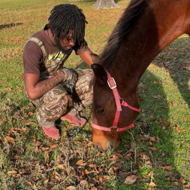 A man with dreadlocks crouching next to a brown horse with a pink halter in a grassy area with fallen leaves.