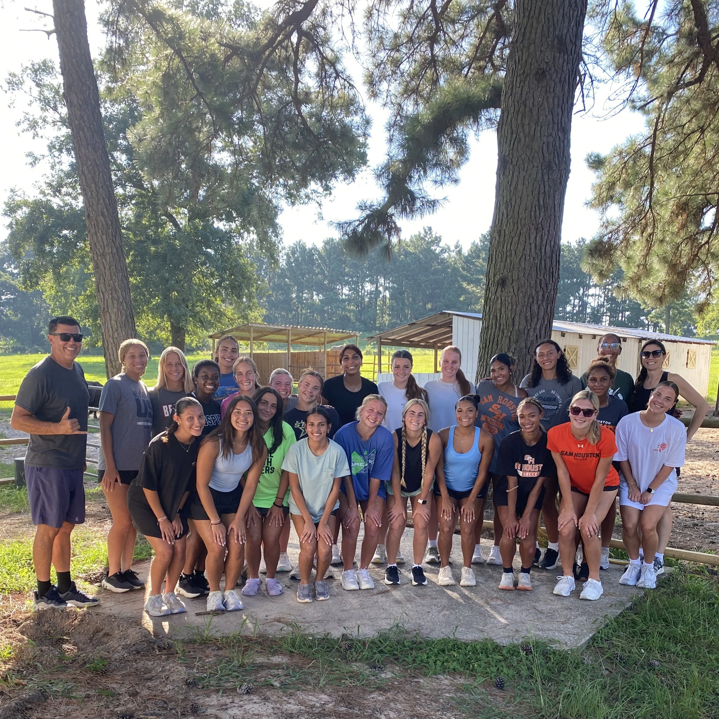 Group of young women and men smiling outdoors, standing on a dirt path with trees and a shed in the background.
