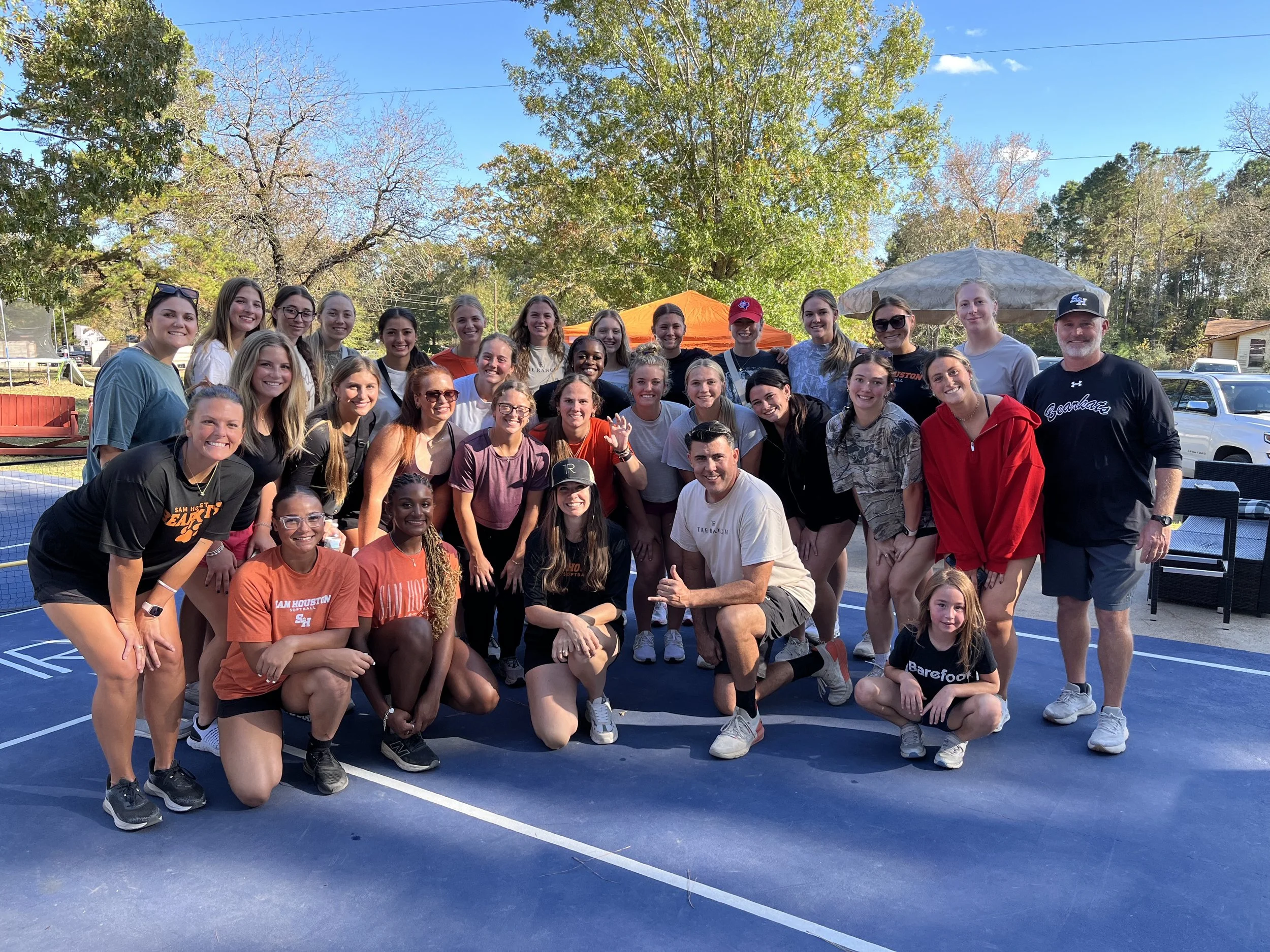 Group of young women and a man posing outdoors on a tennis court with trees and blue sky in the background.