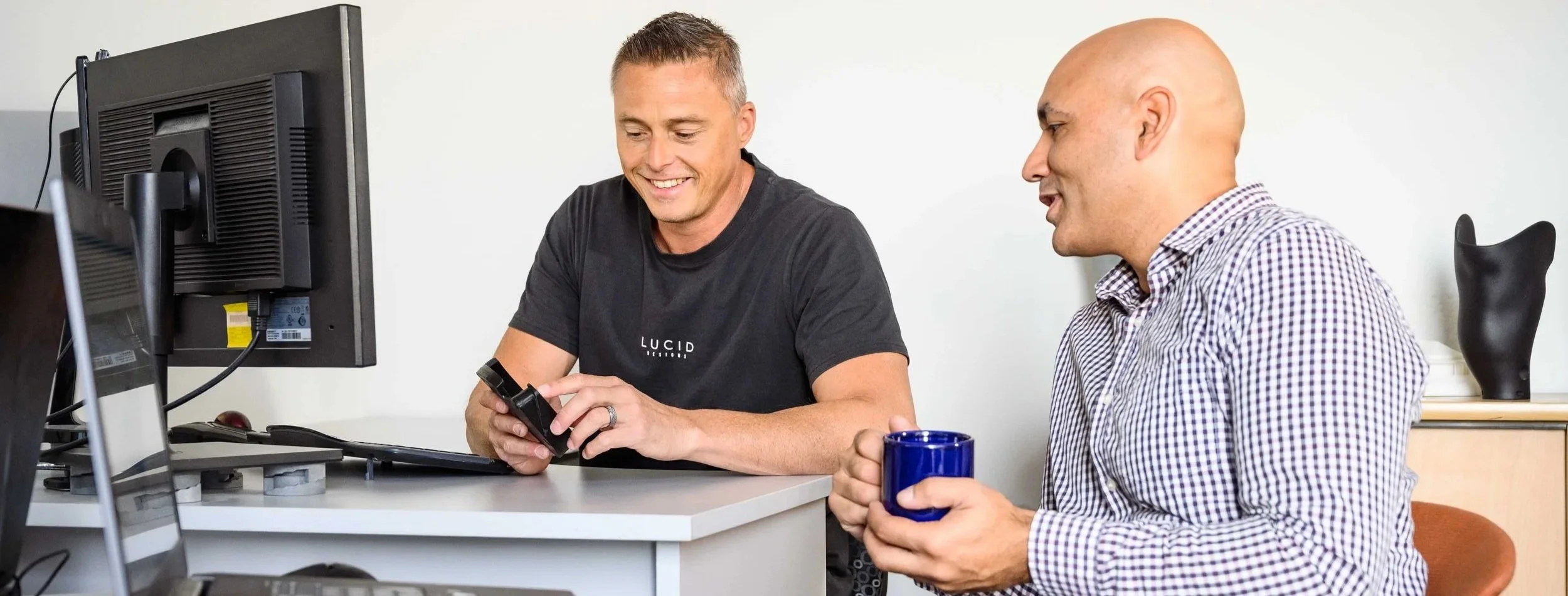 Two men smiling at a desk in an office; one is showing something on a handheld device, while the other is holding a blue mug. There are computers and office supplies on the desk.