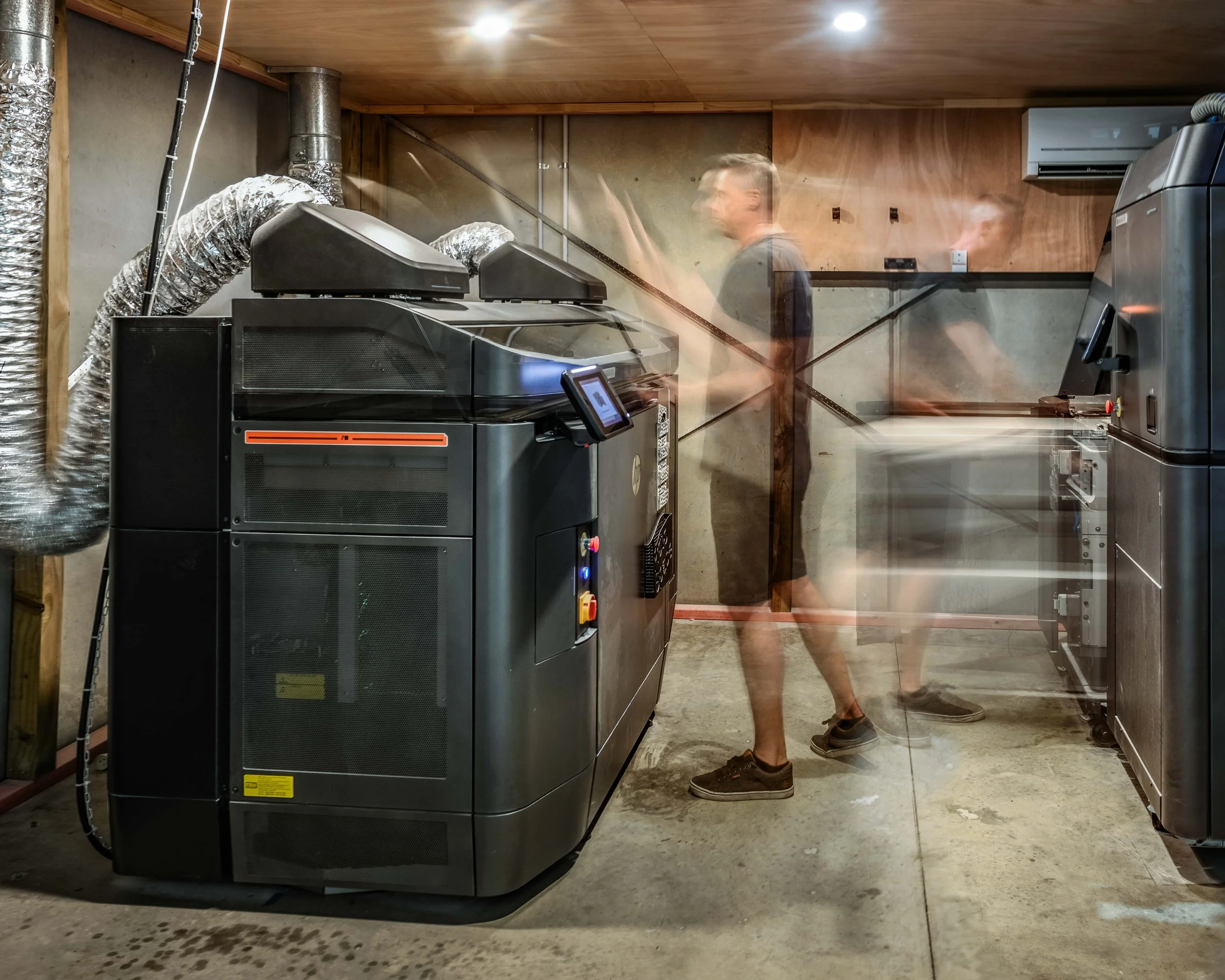 A person standing in a basement laundry room near industrial dryers. The image shows a blurred person wearing dark shorts and a t-shirt, with a concrete floor, wooden ceiling, and ductwork.