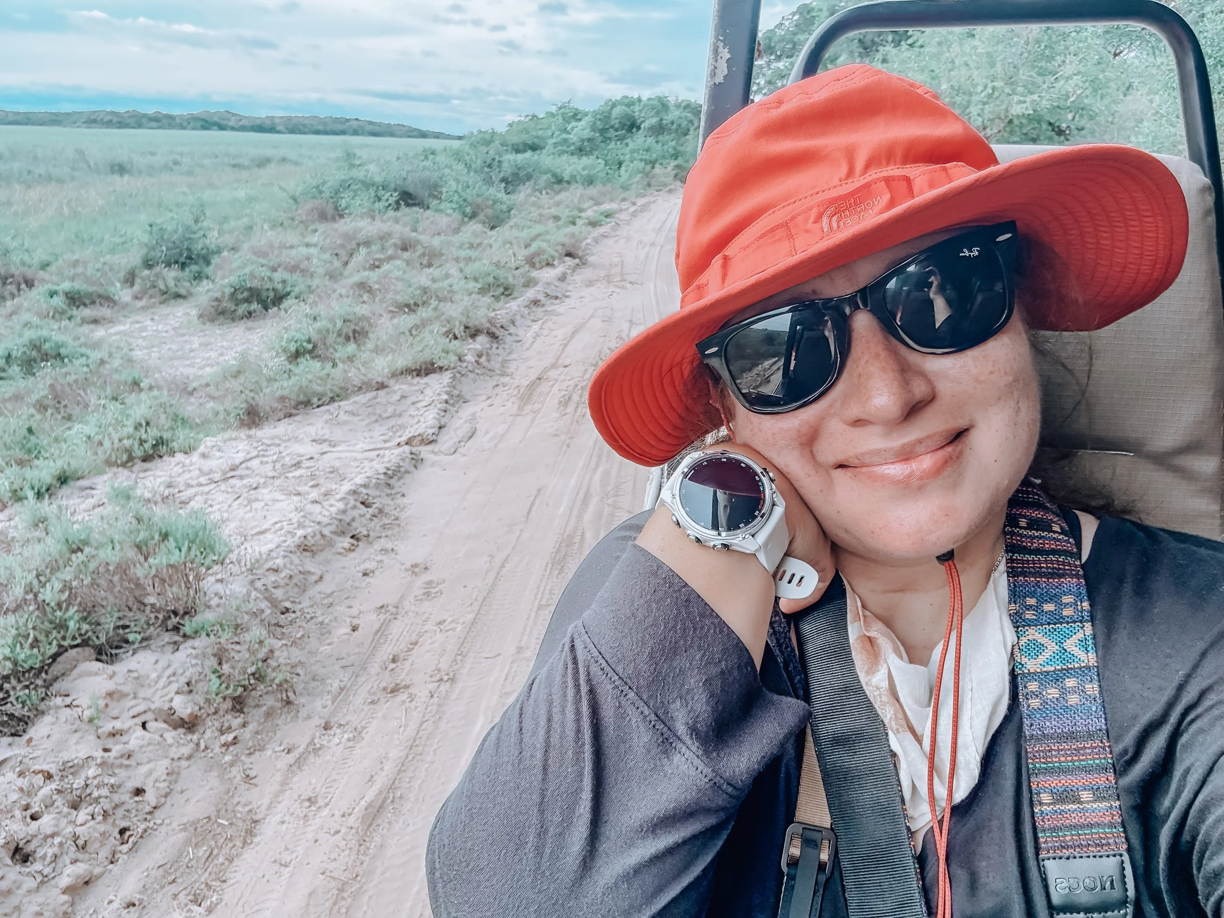 Close-up of a person reclining on a chair outdoors, wearing an orange hat, black sunglasses, a grey jacket, a white watch, and a name tag, with a dirt trail and green fields in the background.