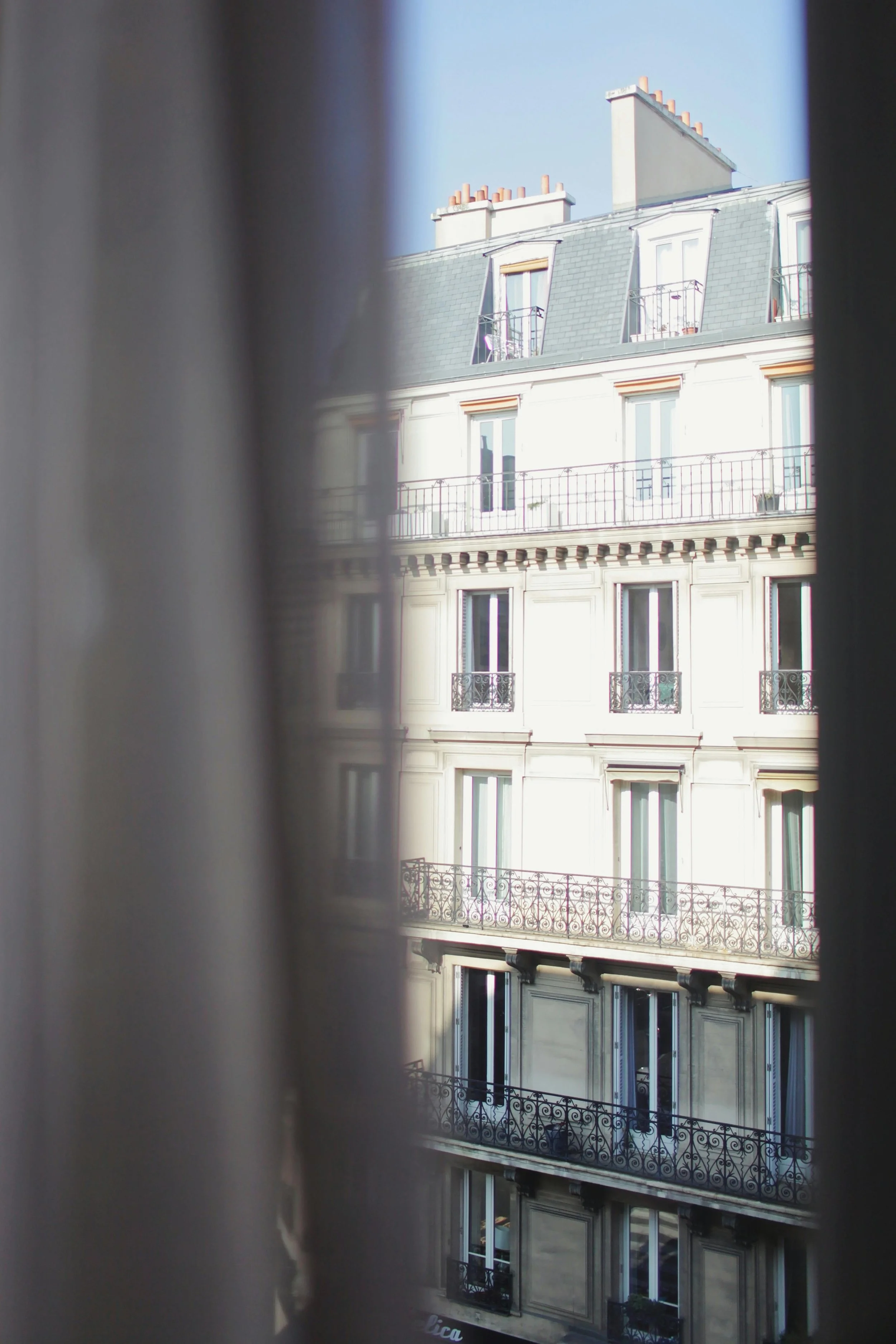 Photo of the upper floors of a classic European-style building through a partially closed window with curtains. The building has several windows and balconies with ornate black iron railings.