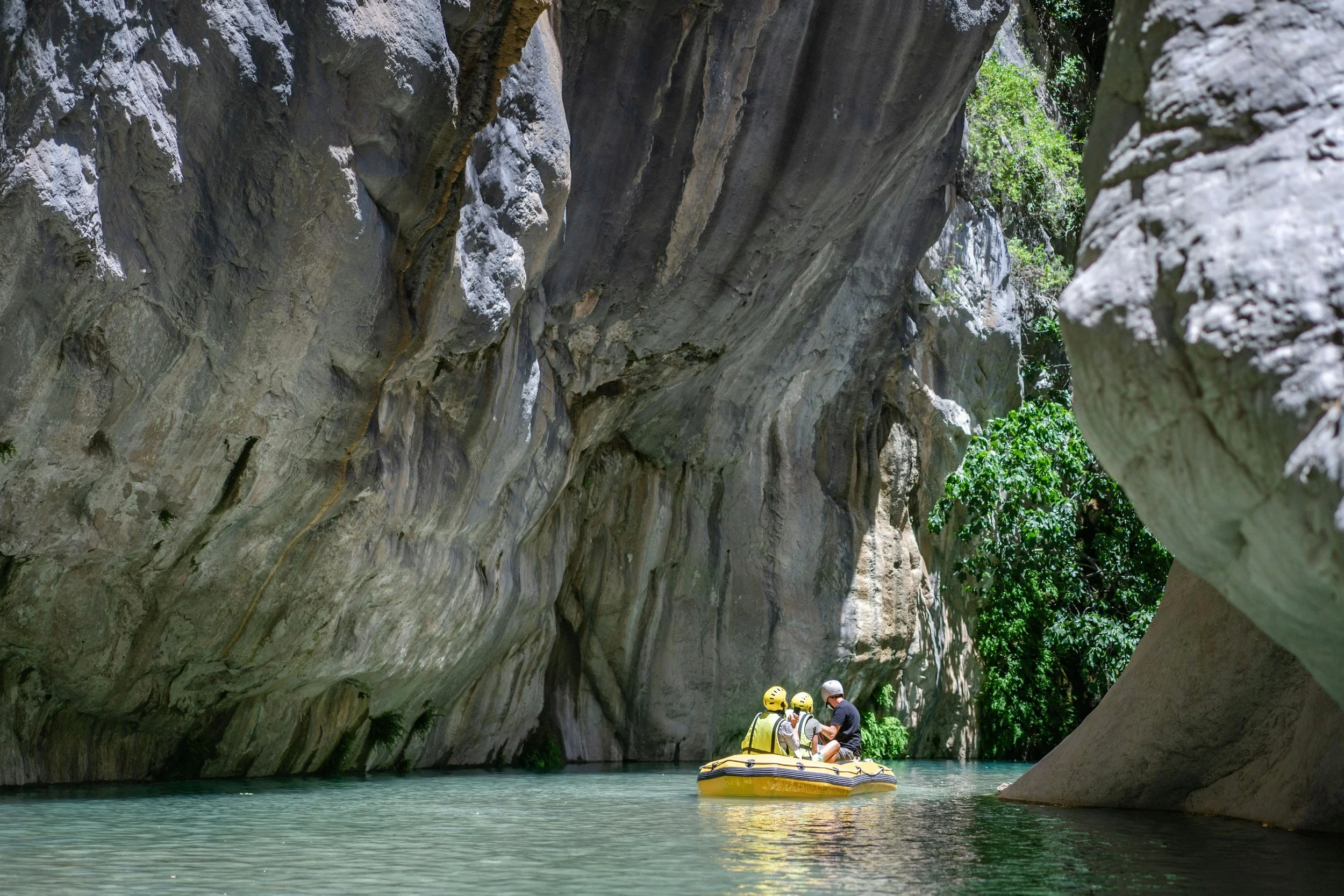 A group of people in yellow helmets on a yellow inflatable boat navigating through a narrow canyon with high, rocky walls and turquoise water.