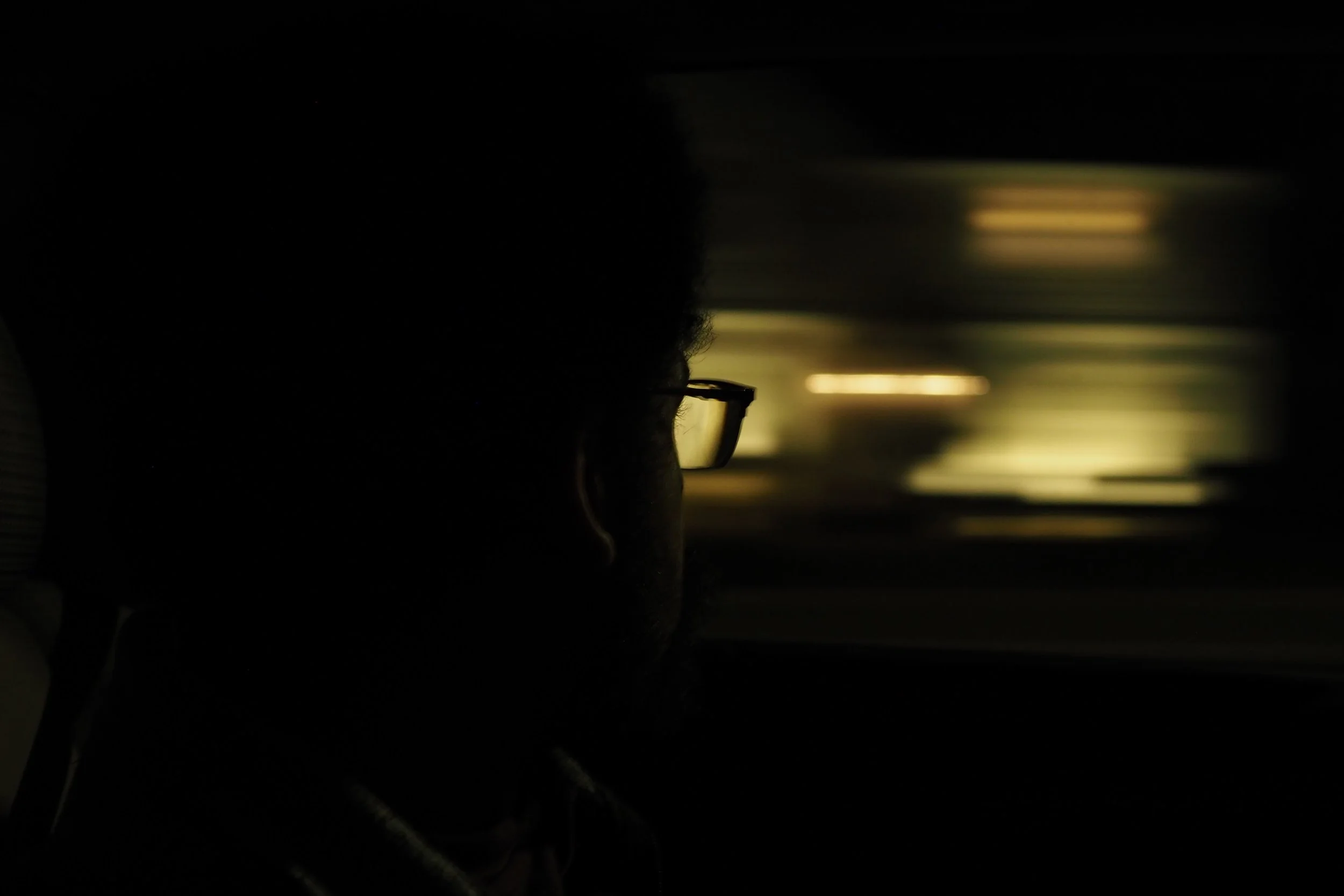 Inside a moving car, the night scene blurs behind the subject. Looking onward, a man with glasses stares into the distance.
