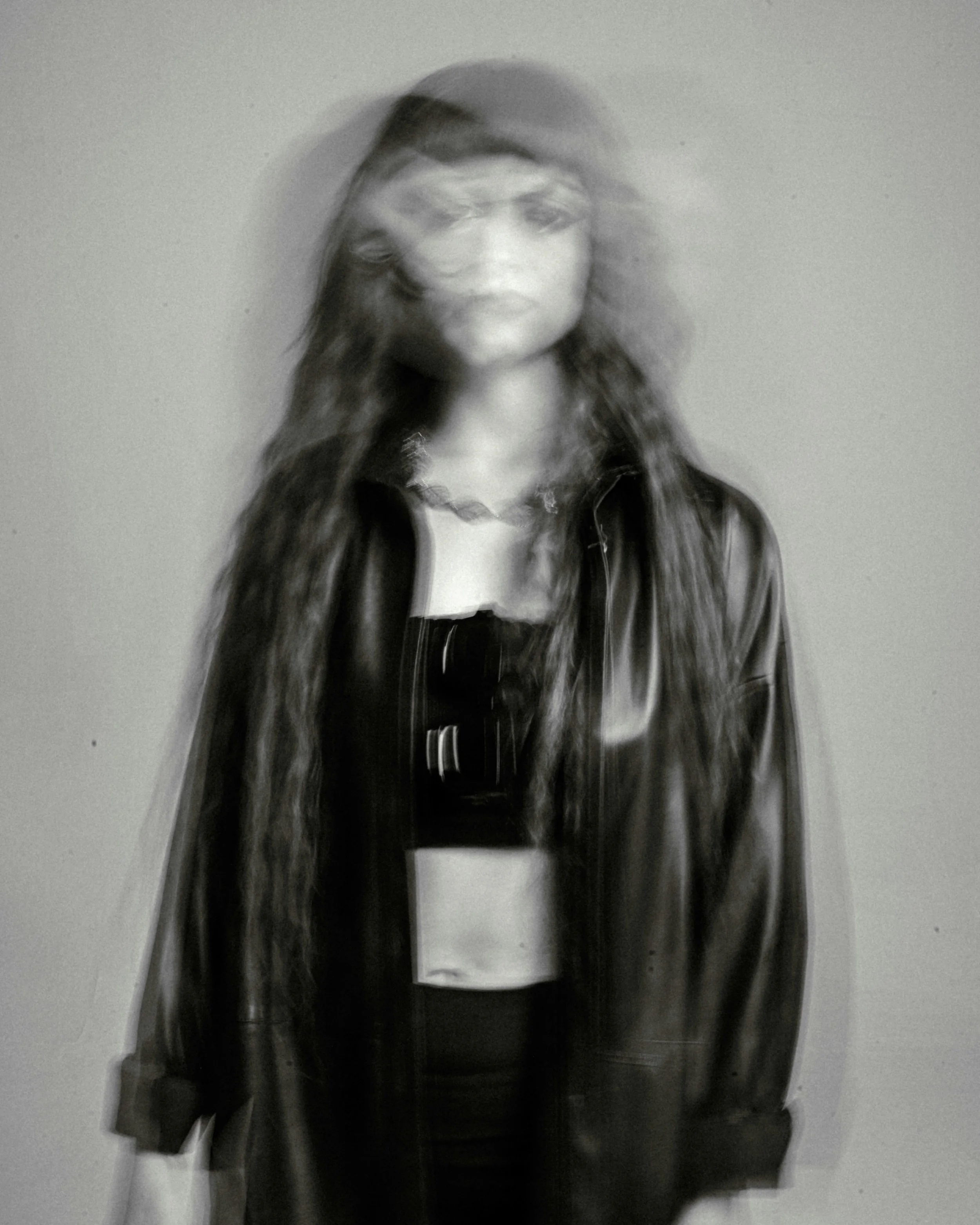 Blurry black and white photo of a young woman with long hair wearing a leather jacket, standing against a plain wall.