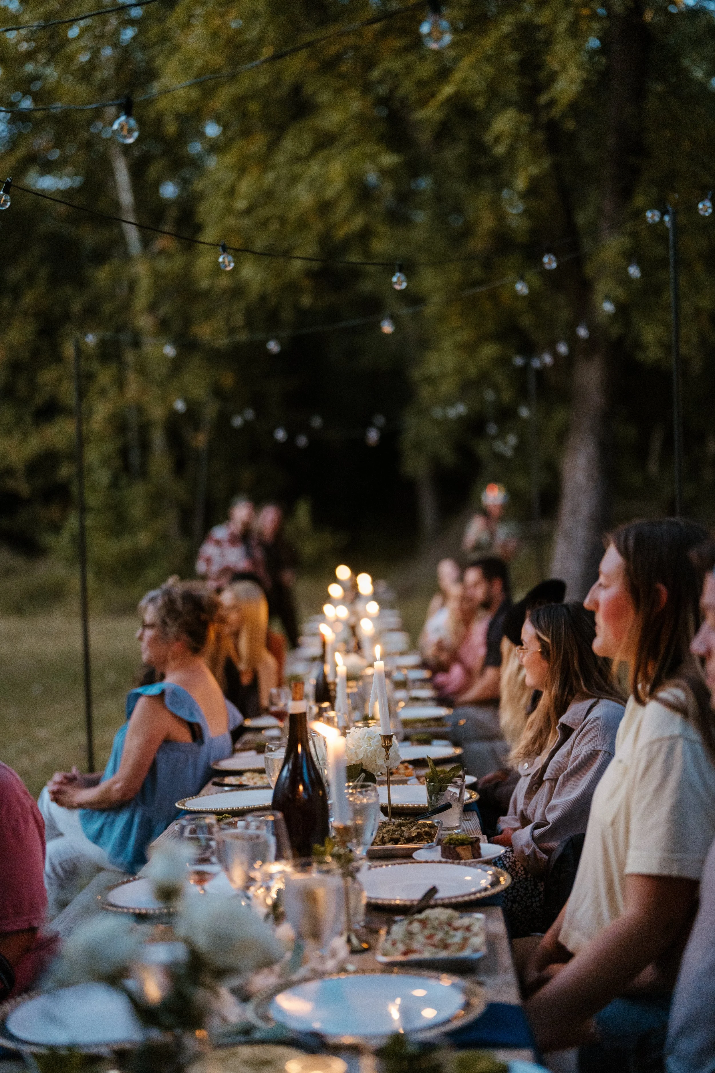 People sitting at a long outdoor dinner table with candles, wine, and food, during dusk or evening, in a wooded area with string lights overhead.