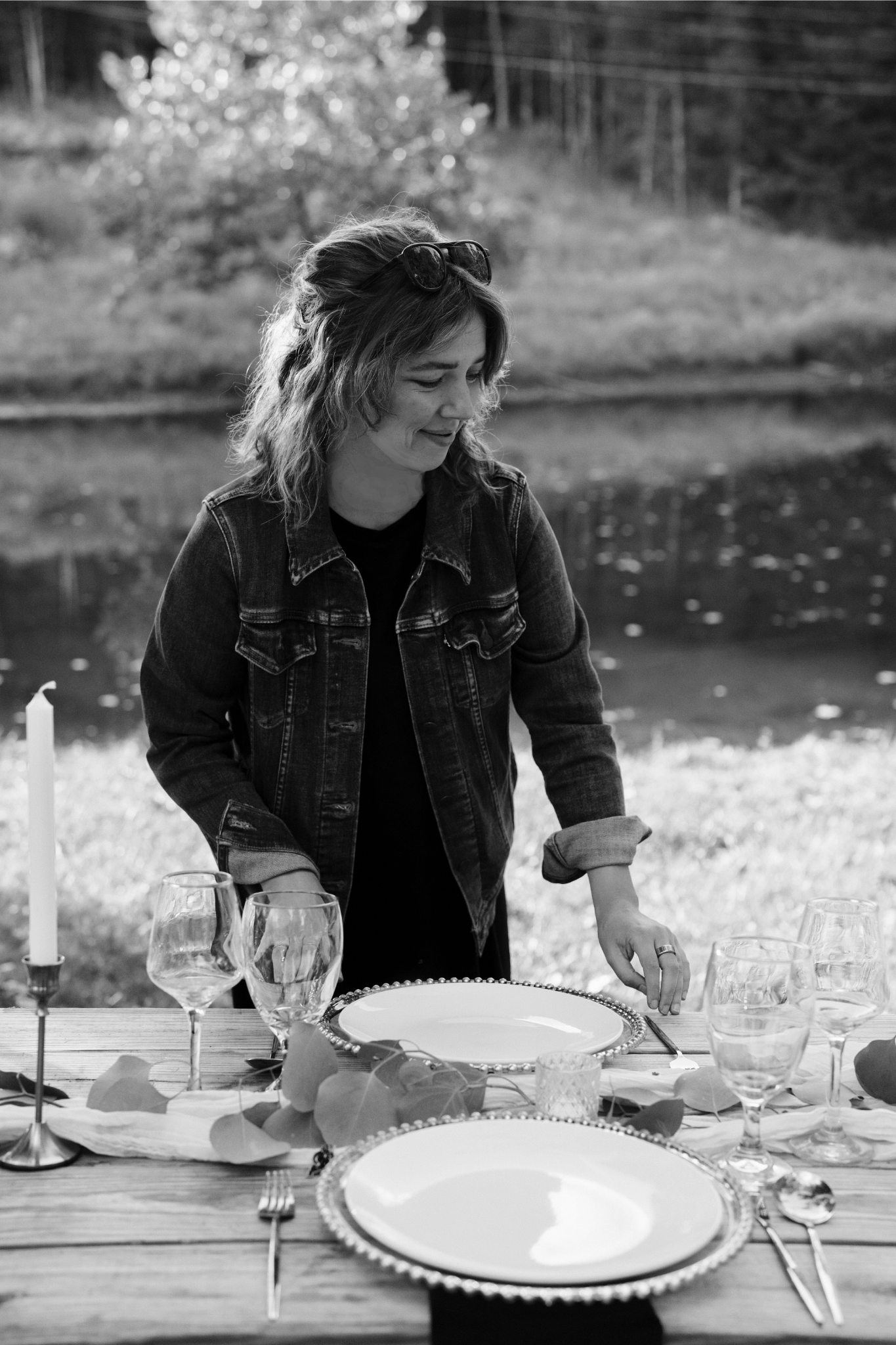 A woman with sunglasses on her head is setting a table outdoors by a lake, with a wooden table and glassware, in black and white.
