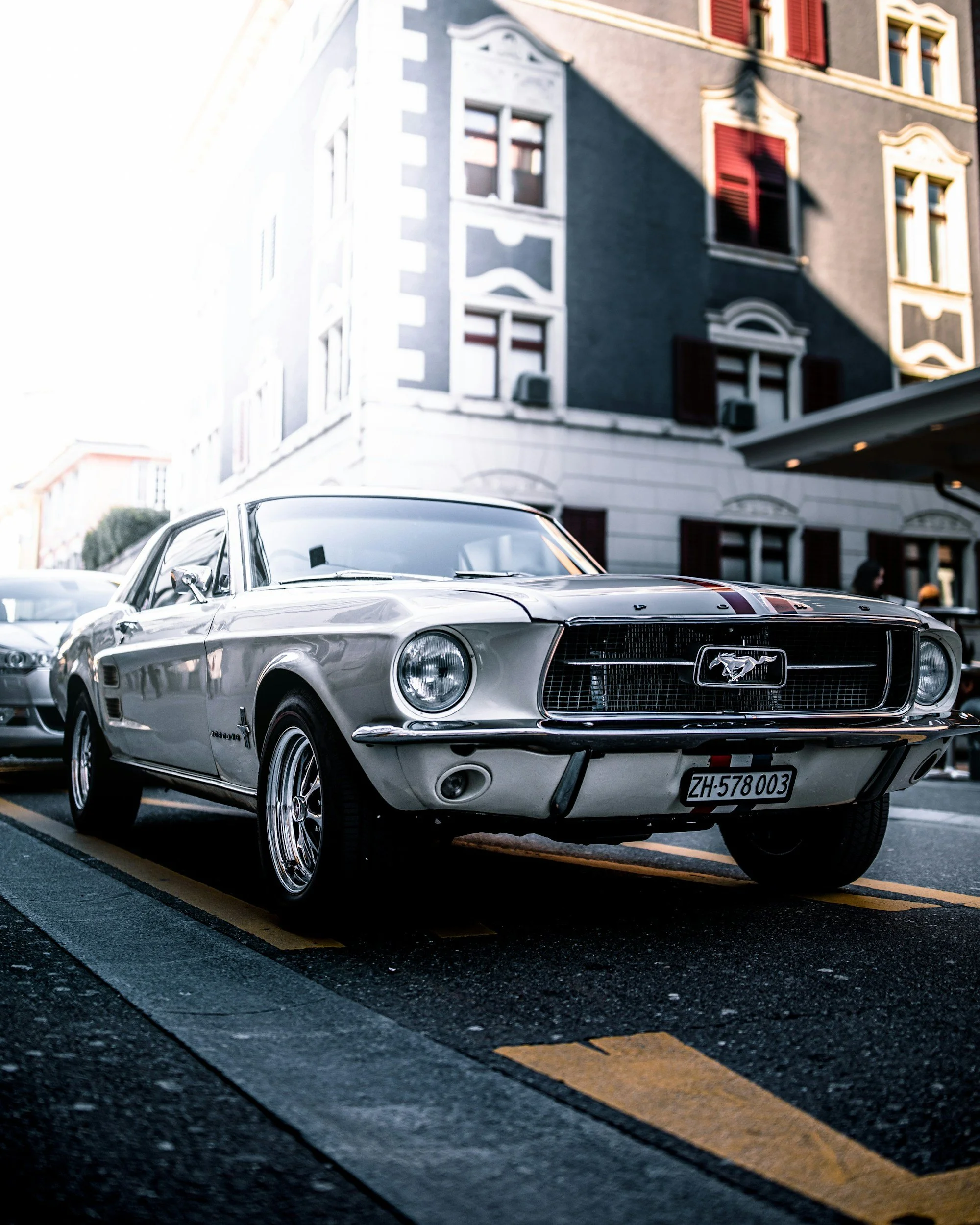 A classic silver Ford Mustang parked on a city street with a building in the background. The building has gray walls with white trim and red shutters. The photo is taken during daylight, and the car is positioned in a parking spot marked by yellow and black lines.