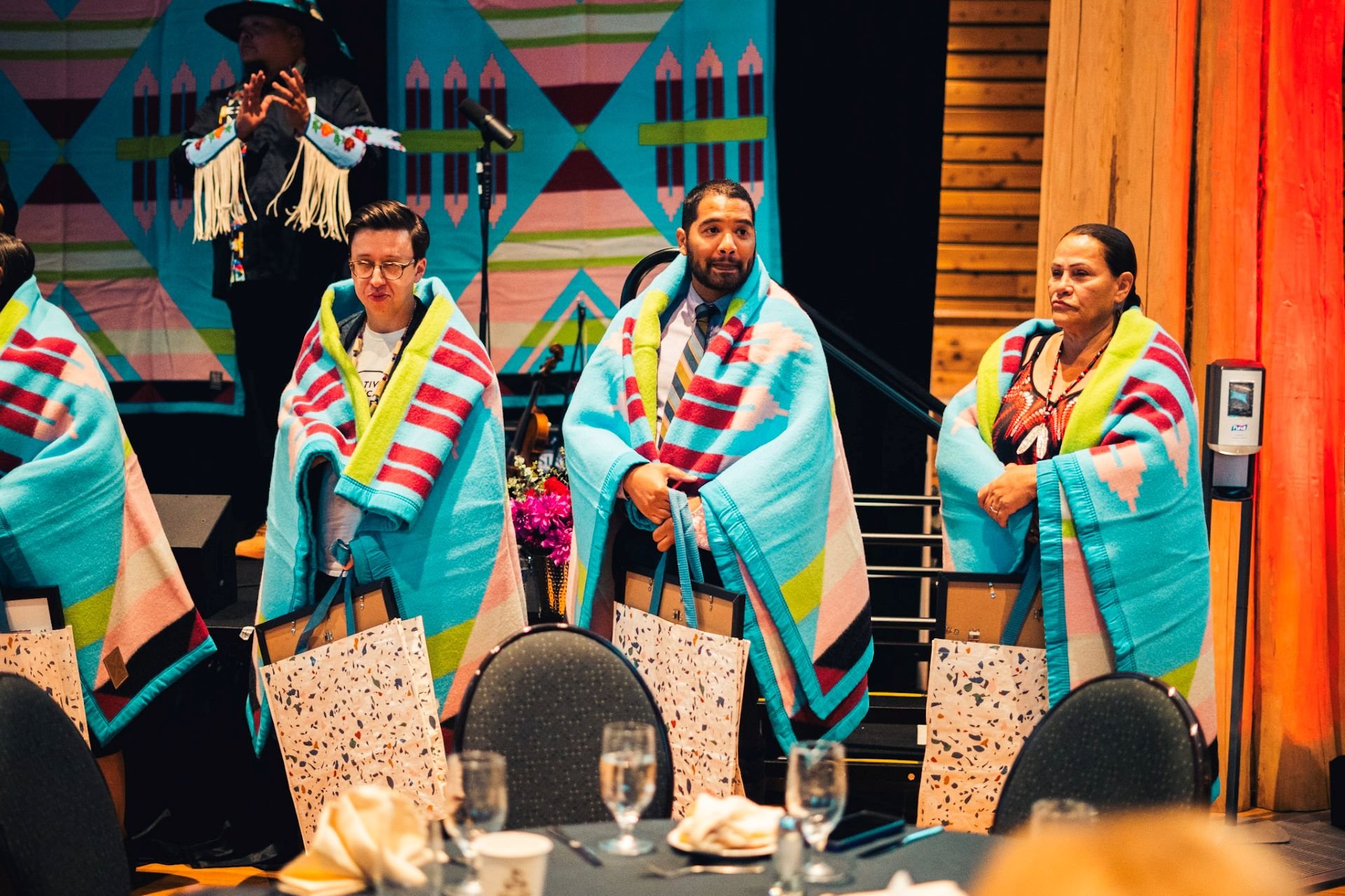 People gathered at an event, wearing traditional blankets, with one standing and gesturing. A woman dressed in Apache attire is on stage, and a colorful quilt hangs in the background.