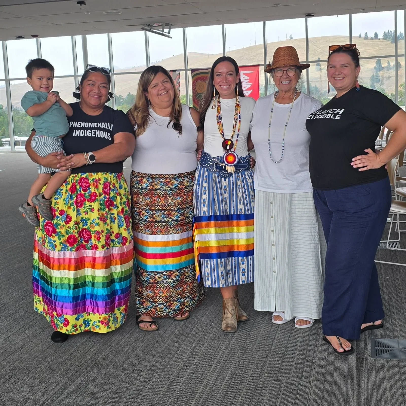Group of six women and one child standing together indoors with mountains visible through large windows in the background. The women are dressed in colorful clothing and smiling at the camera.