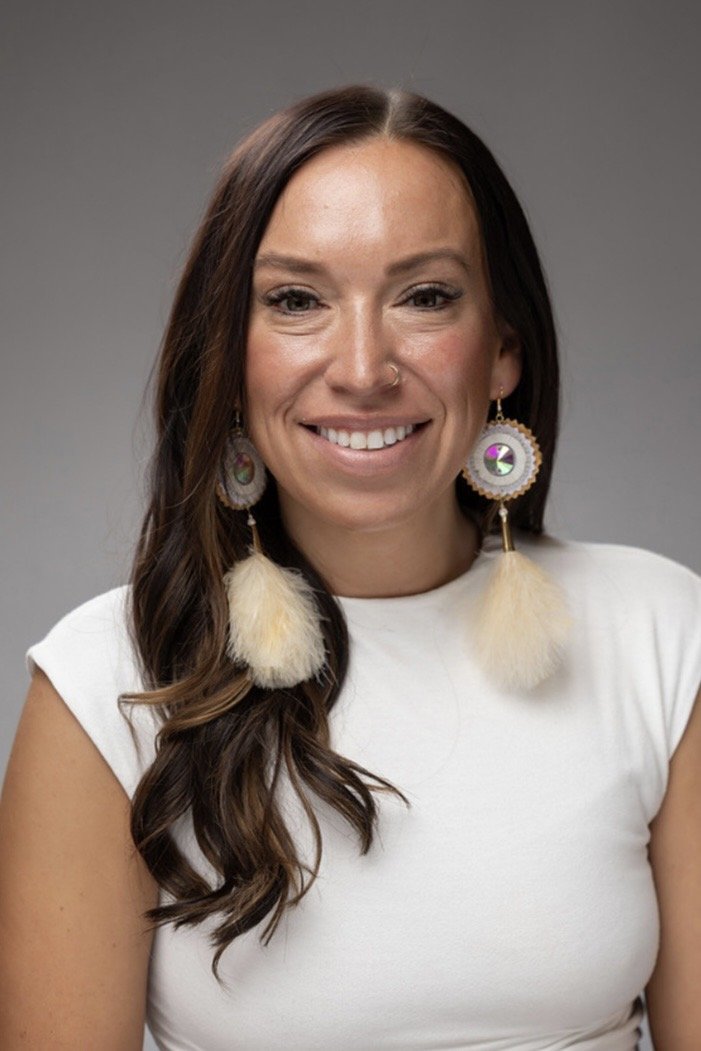 A woman with long brown hair and fair skin is smiling at the camera. She is wearing a white top, hoop earrings with large fluffy feathers, and has a nose ring.