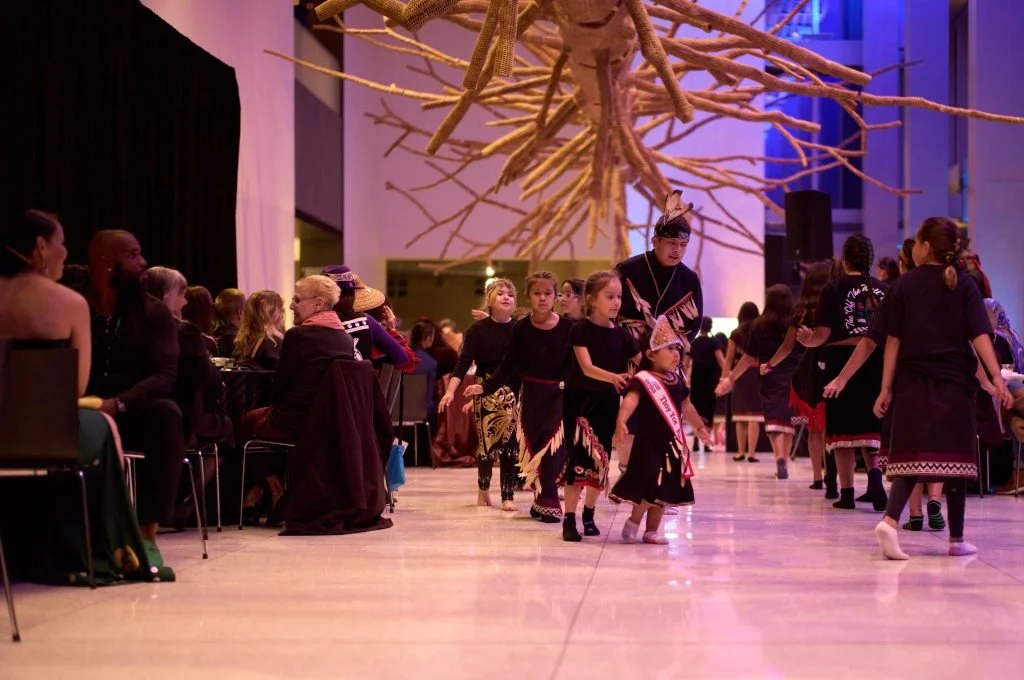 Group of children in traditional indigenous attire performing a dance on stage while audience members watch from tables to the side, in a decorated event space.