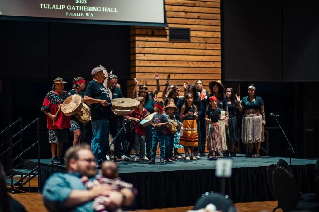 A group of people performing a cultural dance on stage at the 2023 Tulalip Gathering Hall in Tulalip, Washington, with some playing drums and others dressed in traditional attire.