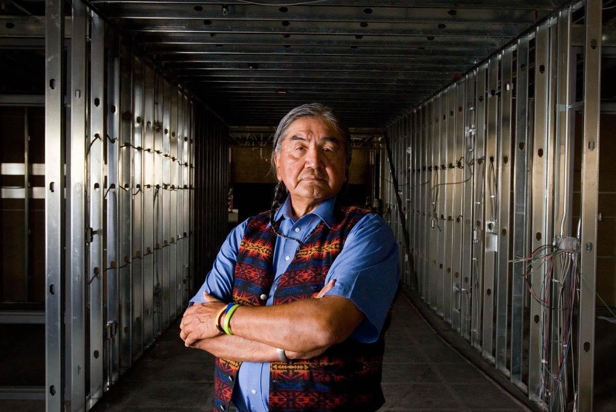 An older man with gray hair and traditional patterned vest standing with arms crossed in a partially constructed building with exposed metal framing.