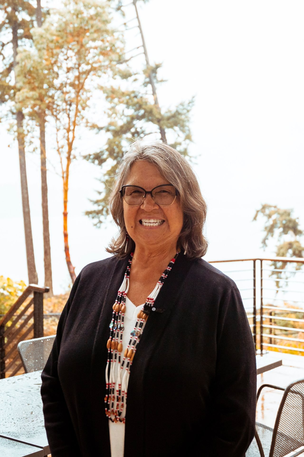 A smiling woman wearing glasses, a black jacket, and a beaded necklace standing on an outdoor patio with trees and a railing in the background.