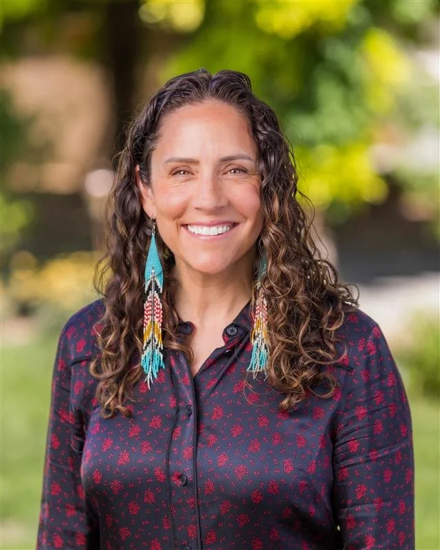 A woman with curly brown hair smiling outdoors, wearing a dark button-up shirt with small red pattern and colorful beaded earrings, with a blurred green and yellow background.