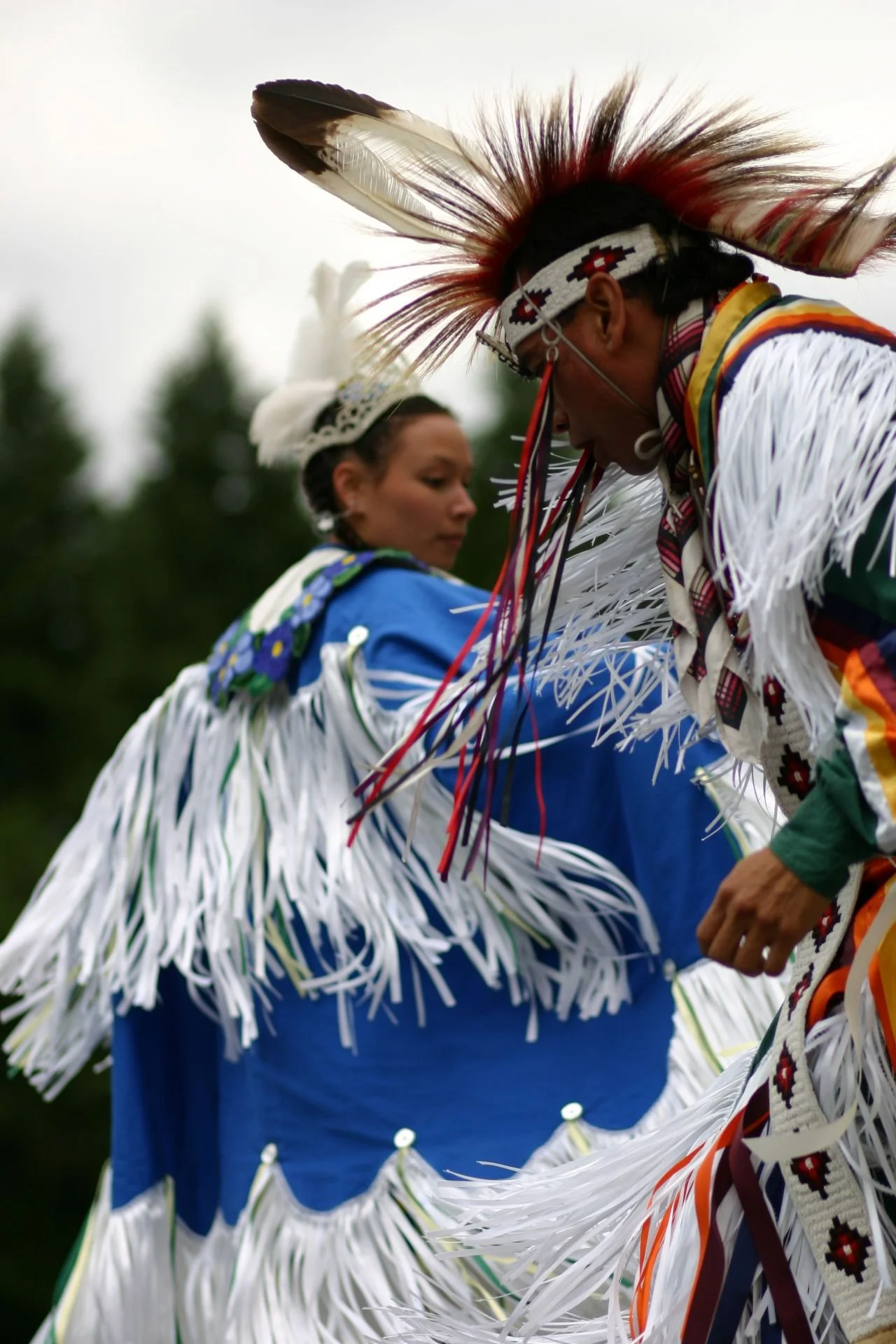 Two Native American dancers wearing traditional regalia, including feathered headdresses and fringed clothing, participating in a cultural dance outdoors.