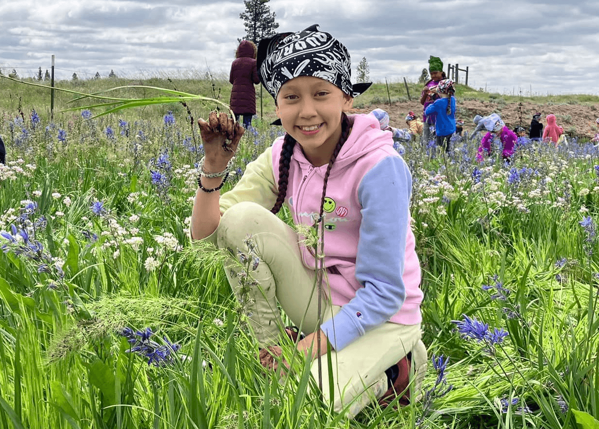 A young girl crouching in a field of wildflowers, holding a flower in her hand, smiling at the camera, with several people in colorful clothing in the background under a cloudy sky.