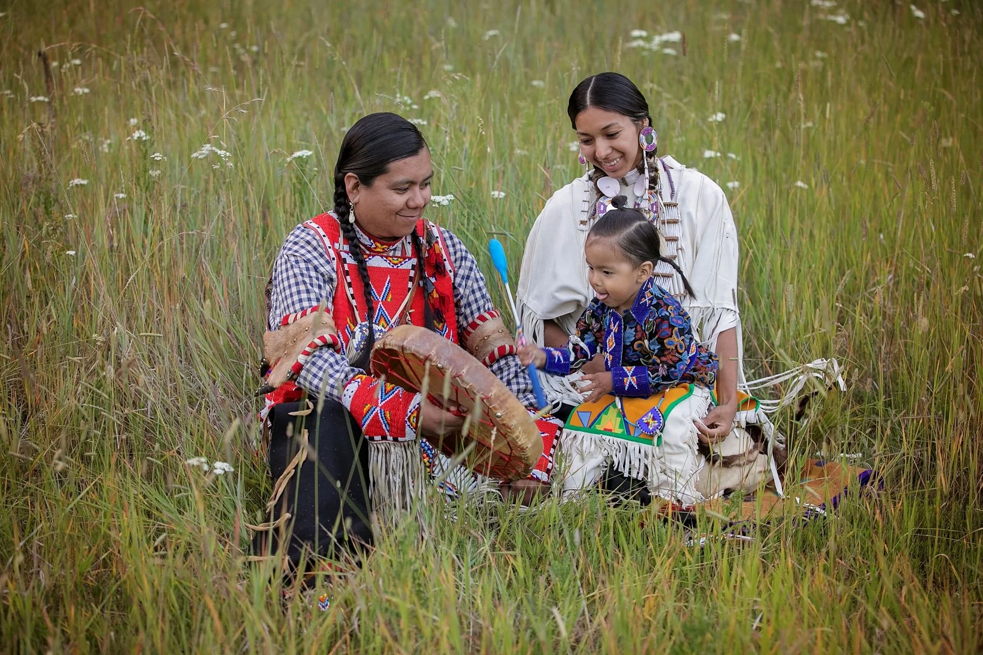 Three Native American women and a girl sitting in tall grass and wildflowers, engaged in a craft activity, wearing colorful traditional clothing and accessories.