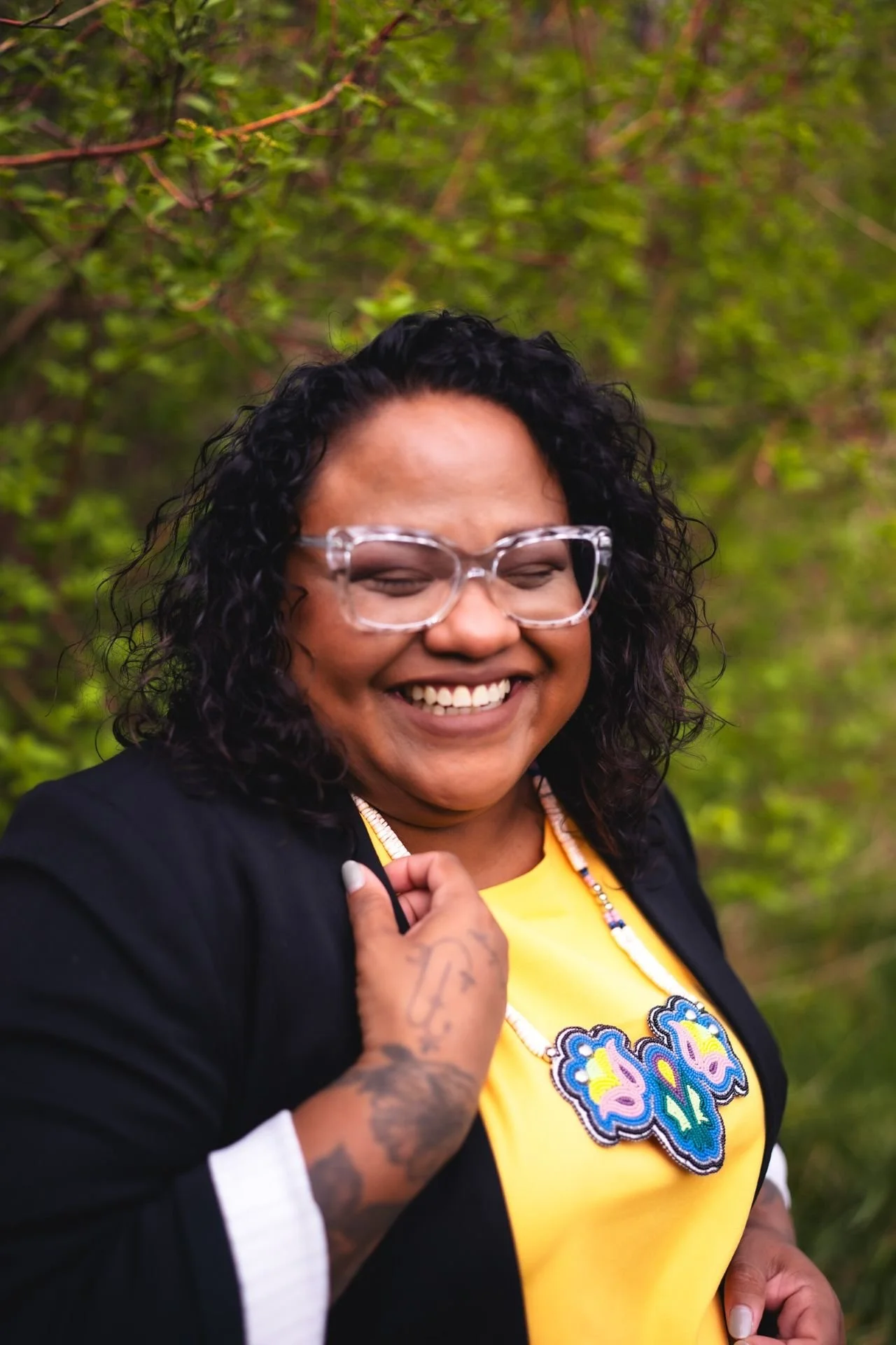 A woman with curly black hair and glasses smiling outdoors, wearing a yellow shirt with a colorful embroidered design, and a black jacket, standing in front of green foliage.