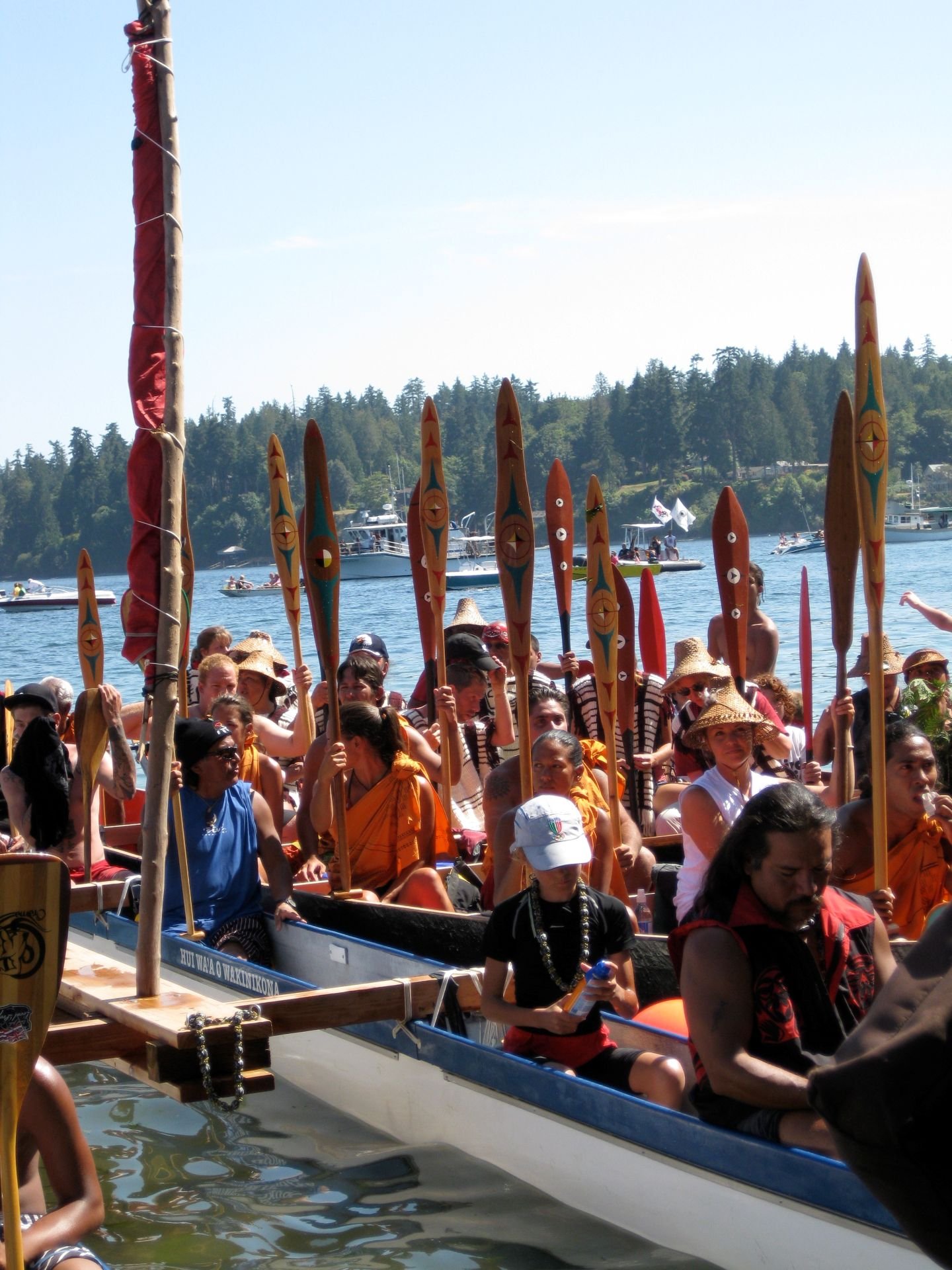 A large group of people in traditional Polynesian attire participating in a cultural festival on the water, with many people holding carved wooden paddles, colorful leis, and wearing straw hats. Several boats and yachts are visible in the background on a sunny day with clear blue skies.