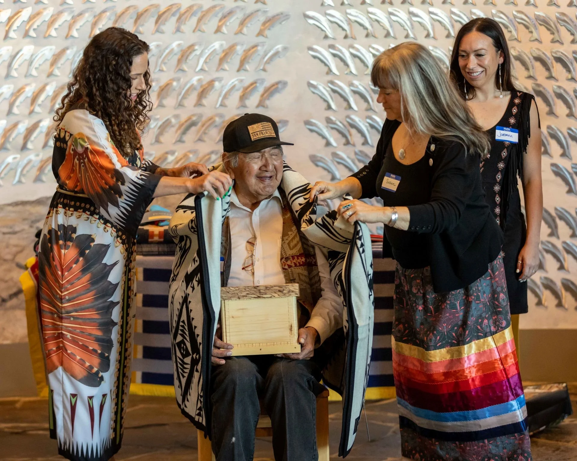 Four women and an elderly man are gathered together, with the elderly man seated in a wheelchair. The women are smiling and appear to be presenting him with a framed item, with a decorative wall behind them featuring numerous silver and gold dolphin-shaped ornaments.