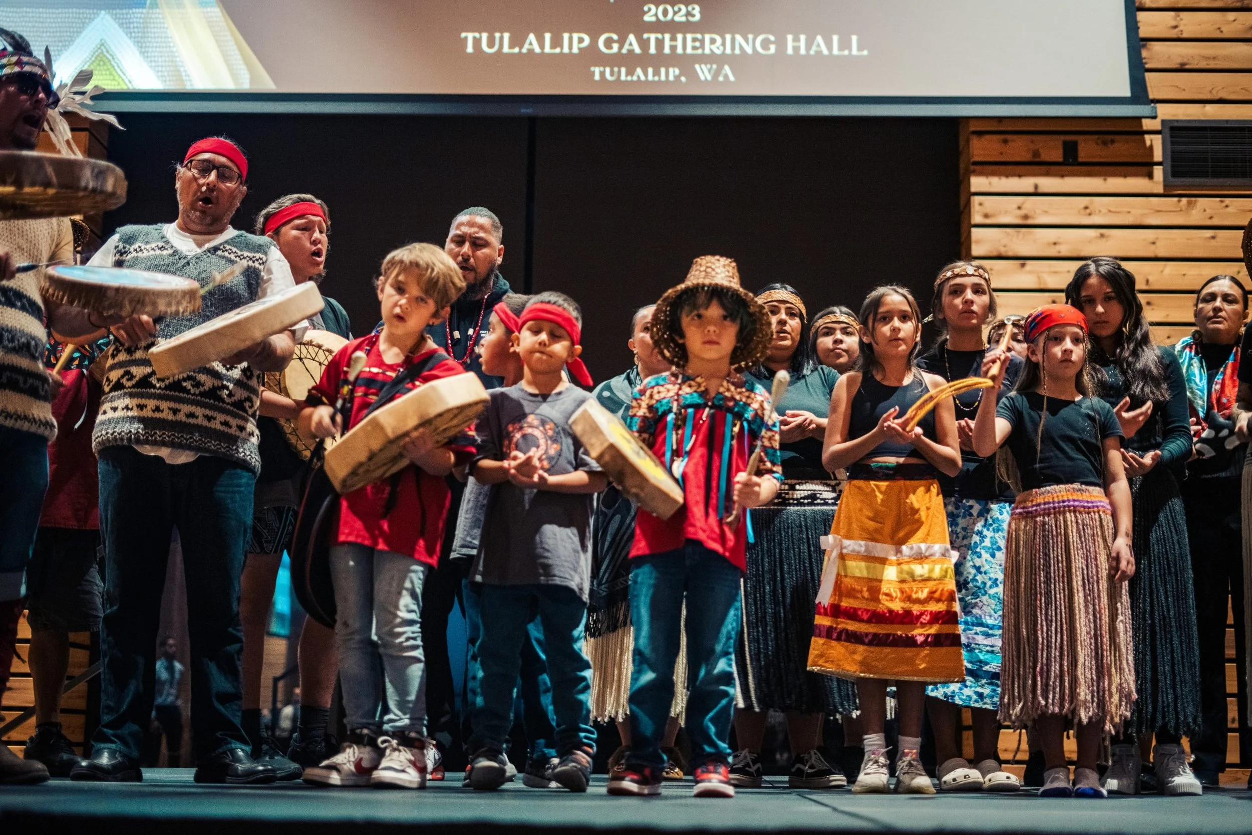 Children and adults performing on stage at Tulalip Gathering Hall during a cultural event, dressed in traditional clothing and playing drums.