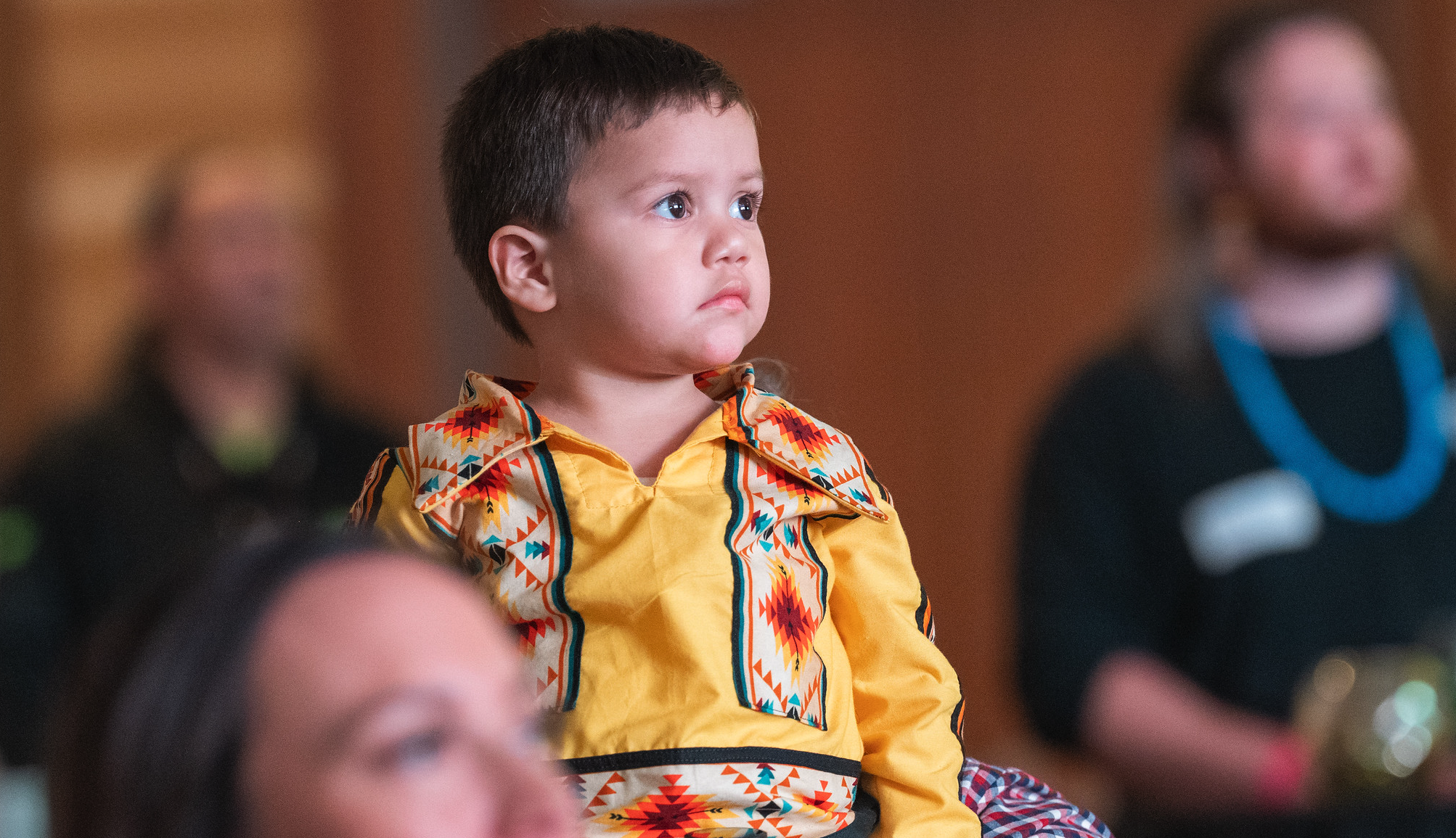 A young boy with short dark hair and light skin, wearing a colorful Aztec-patterned yellow shirt, appears to be attending an event or classroom, sitting attentively.