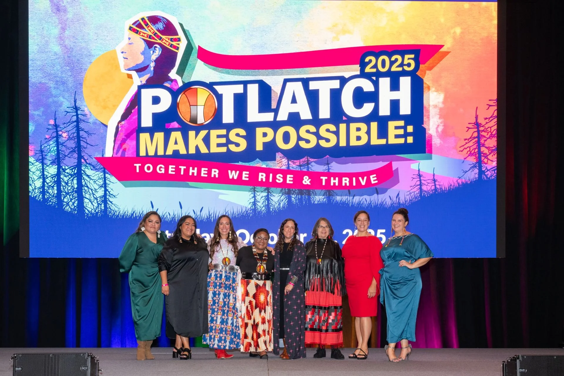 A group of eight women standing on a stage in front of a colorful backdrop that reads 'Potlatch 2025 Makes Possible: Together We Rise & Thrive,' with illustrations of trees, a large yellow circle, and a portrait of a woman wearing traditional Native American attire. The women are dressed in various styles, including traditional and modern clothing.