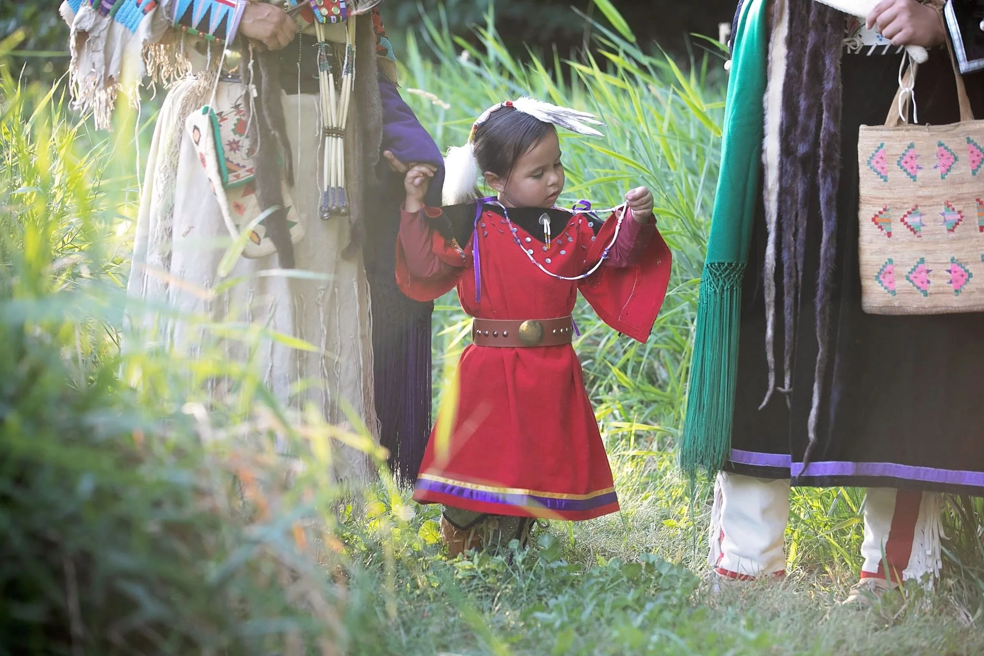 A young girl wearing a red traditional dress with beads and feather headdress stands among tall green grass, holding hands with two adults, and surrounded by people wearing cultural clothing and accessories in an outdoor setting.