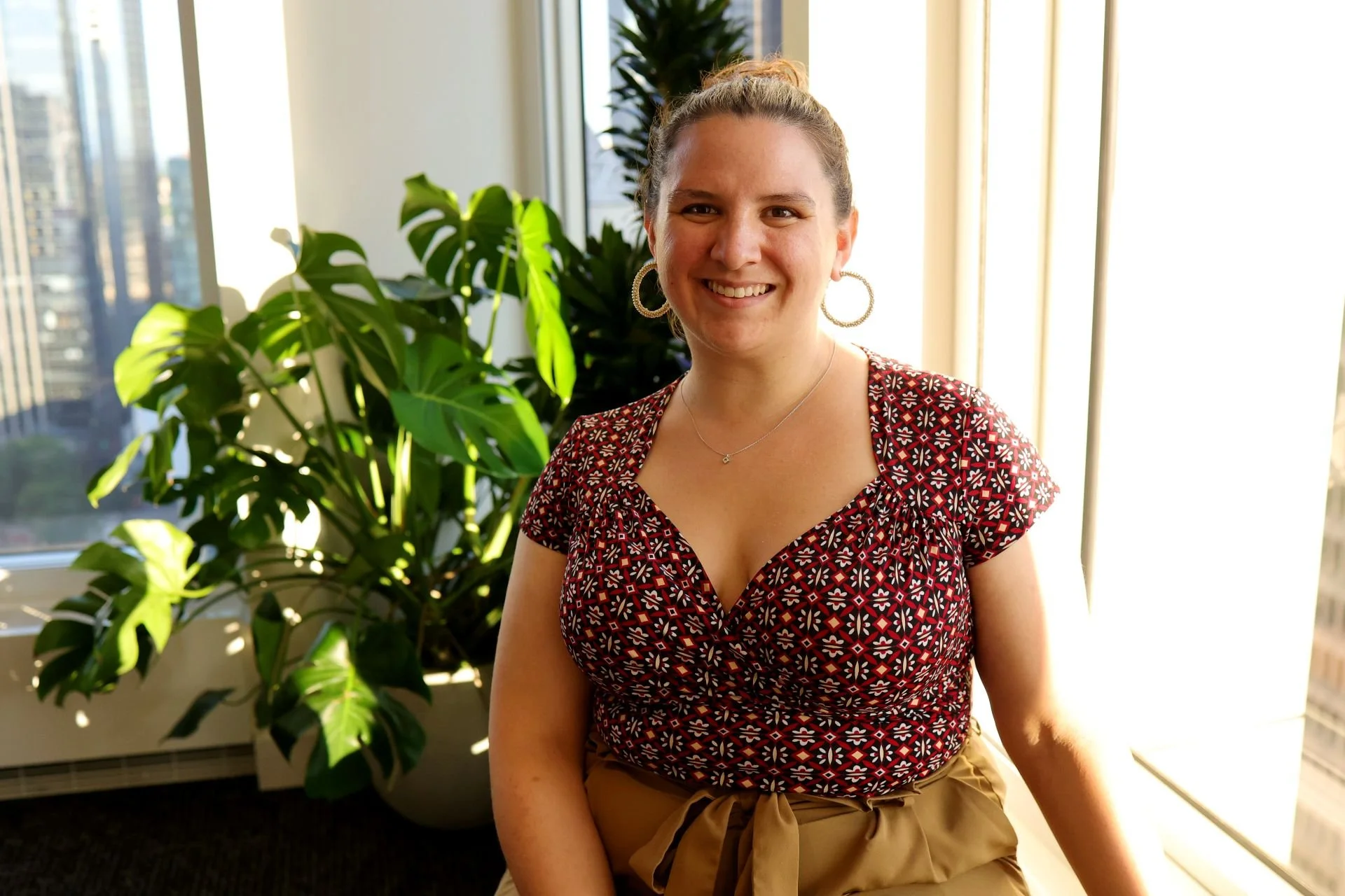Smiling woman with curly hair pulled back, wearing hoop earrings, a patterned red and white top, and beige pants with a bow, standing near a large green plant in front of a window with city buildings visible in the background.