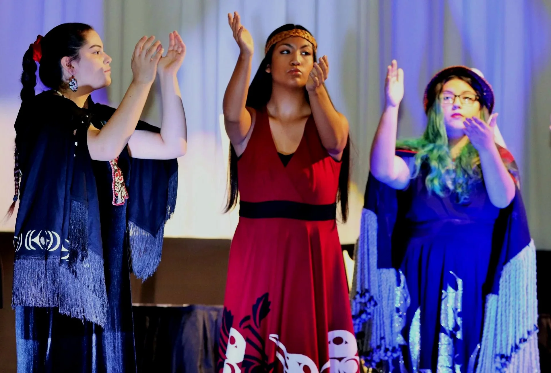 Group of women on stage, wearing colorful attire, with one in a red dress and headband, gesturing with their hands during a performance.