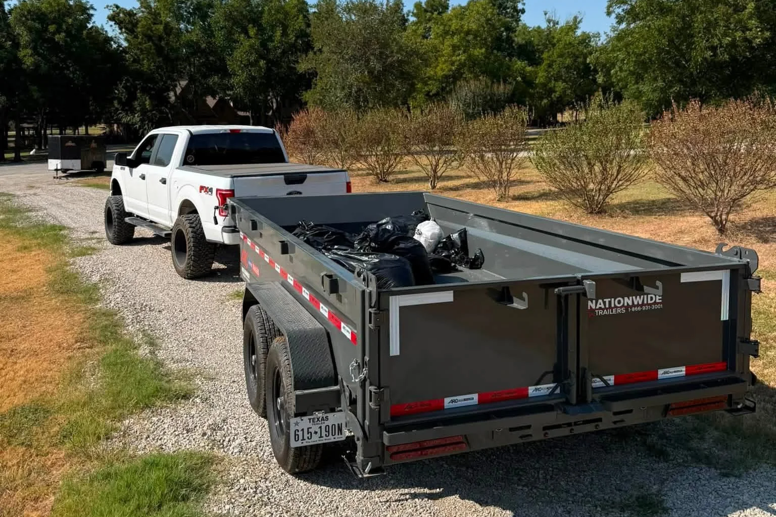 White pickup truck attached to a black utility trailer on a gravel driveway, with trees and bushes in the background.