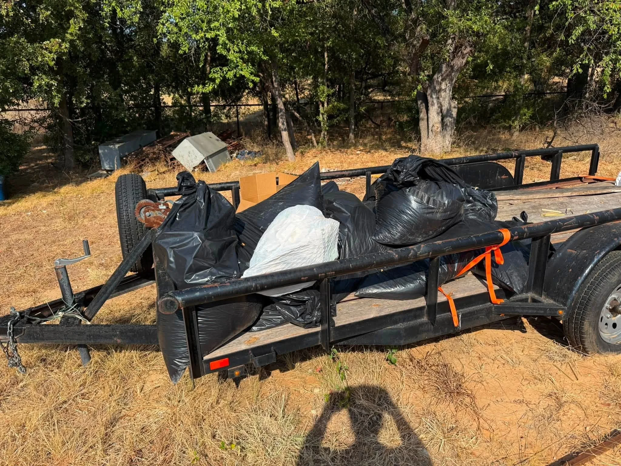 A flatbed utility trailer loaded with black trash bags, a cardboard box, and miscellaneous debris parked on dry grass in front of trees.