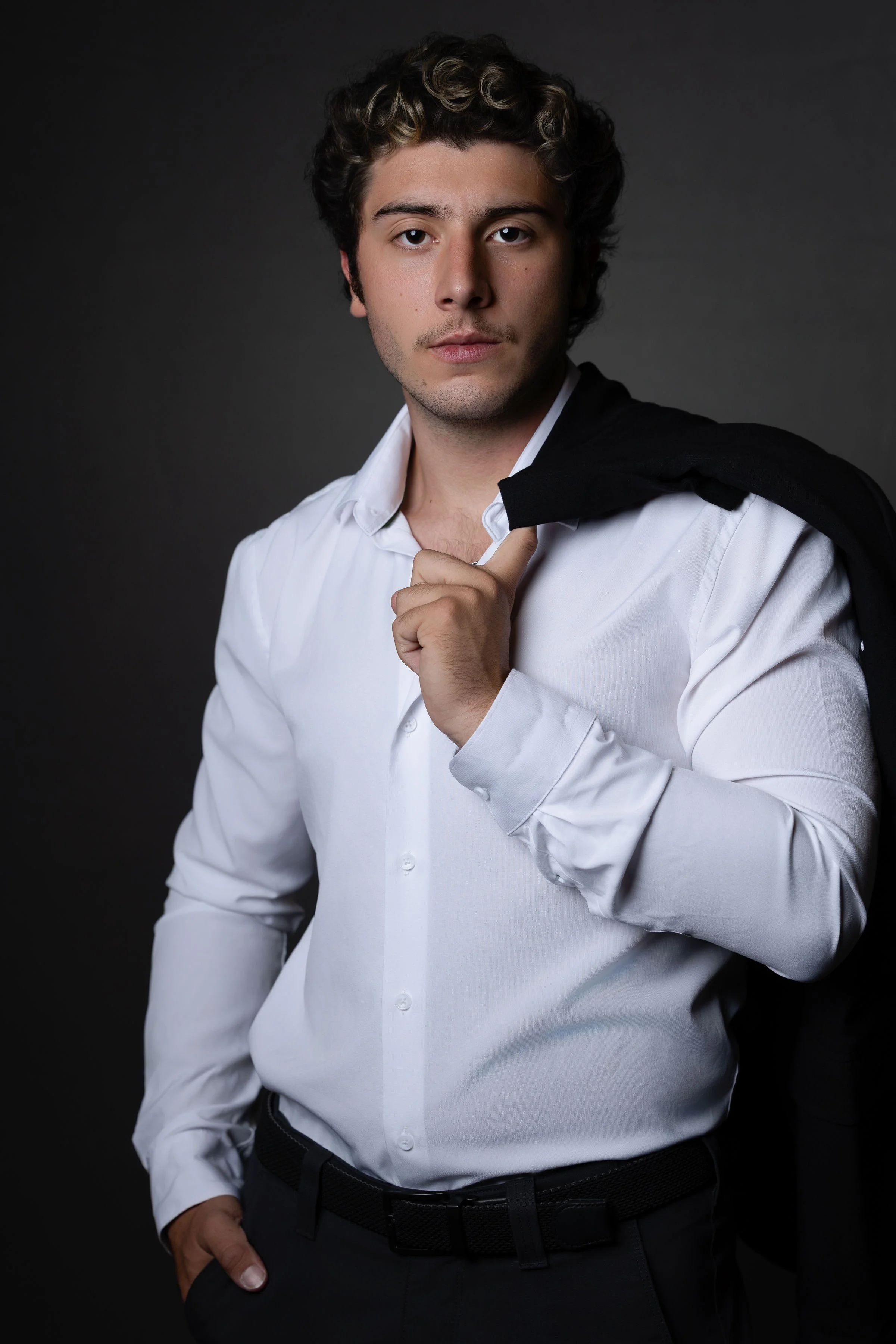 A young man with curly hair, wearing a white dress shirt, holding a black jacket over his shoulder, standing against a dark background.