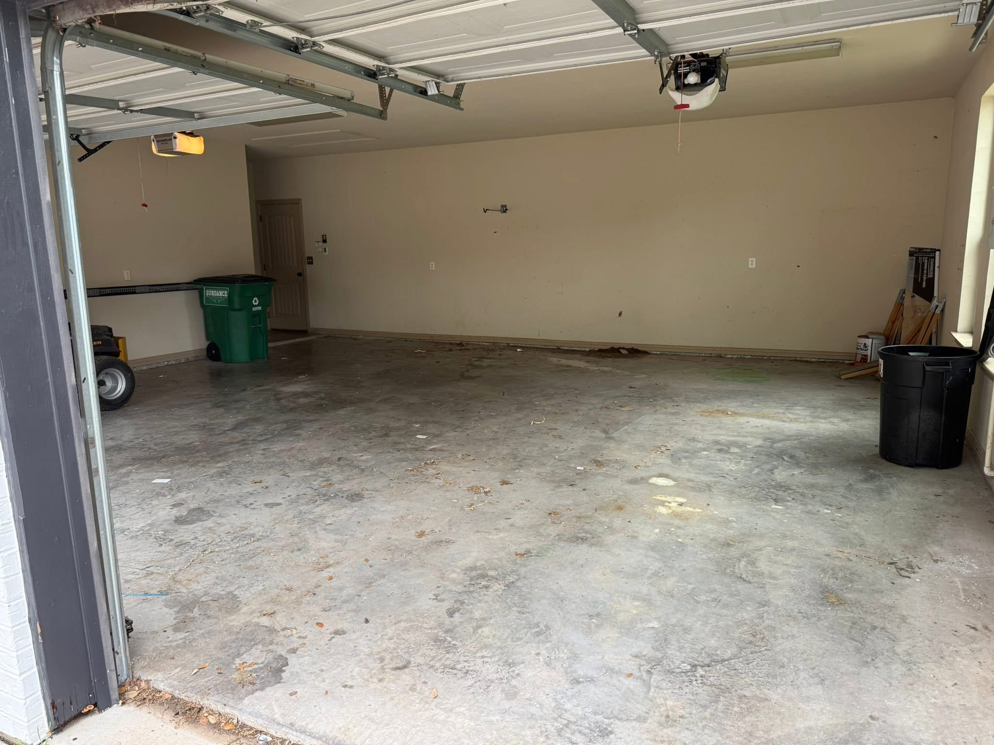 Empty garage with a concrete floor, beige walls, and a partially open metal garage door. There is a green recycling bin, a black trash can, and some construction materials and tools in the garage.