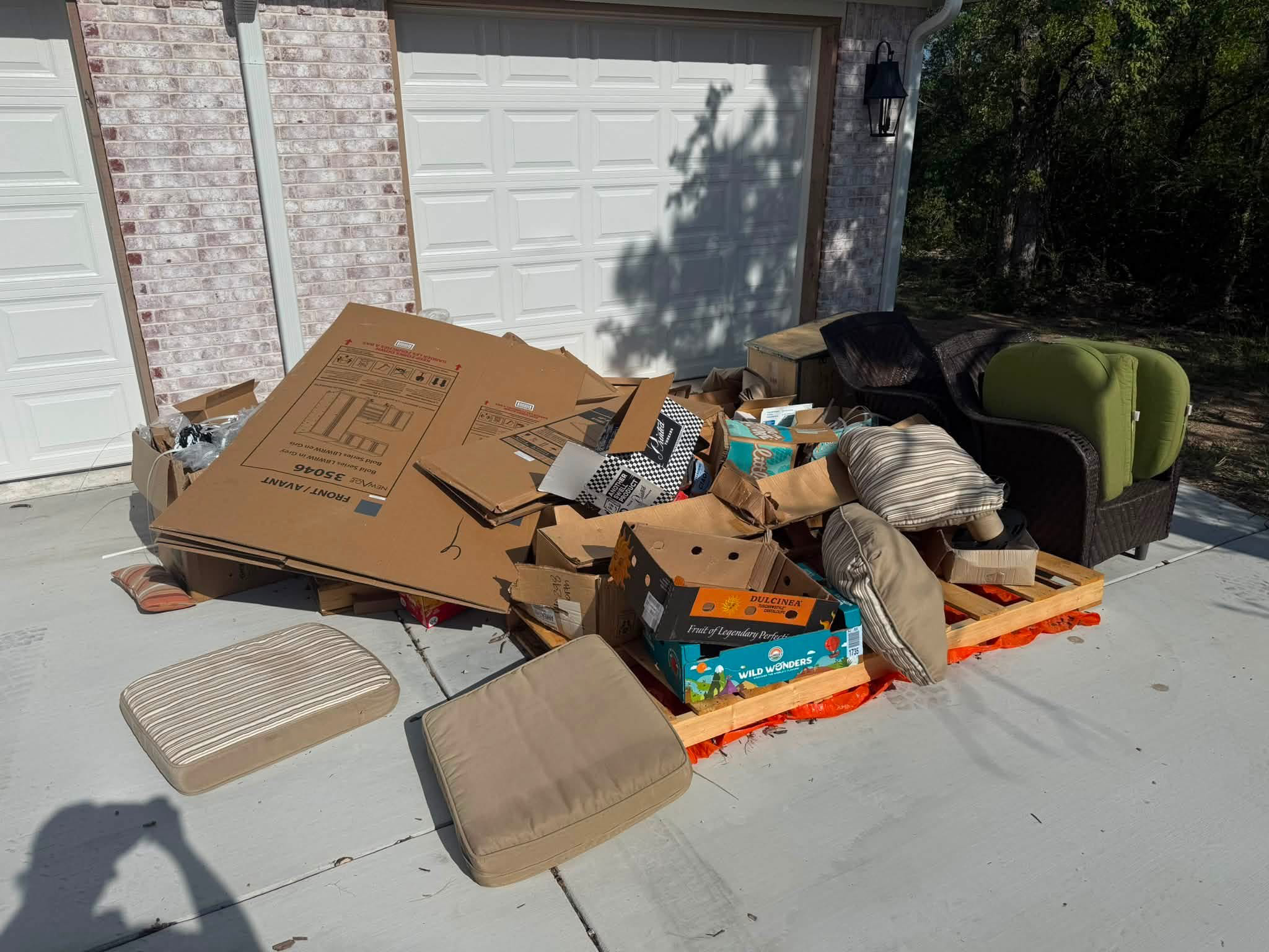 A pile of cardboard boxes, some open and some flat, placed on a wooden pallet on a concrete driveway. There's an outdoor chair, a green cushioned seat, and a loveseat with striped cushions nearby. The background features a brick wall, white garage doors, and green foliage. Shadows are cast on the garage door from nearby trees or bushes.