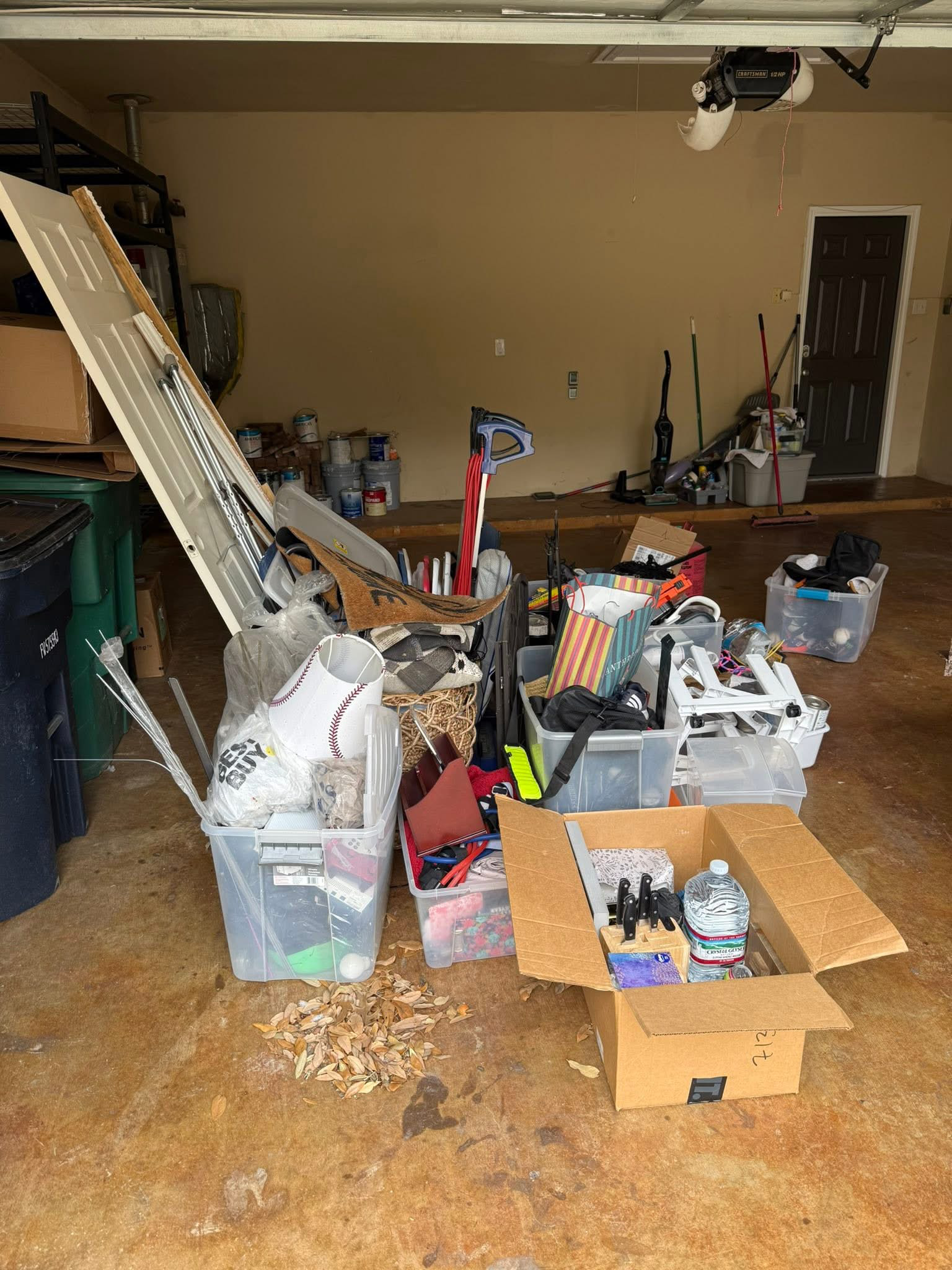 A cluttered garage with various boxes, storage containers, and tools scattered across the floor, against a beige wall and wooden floor, with some paint cans stacked on the side.