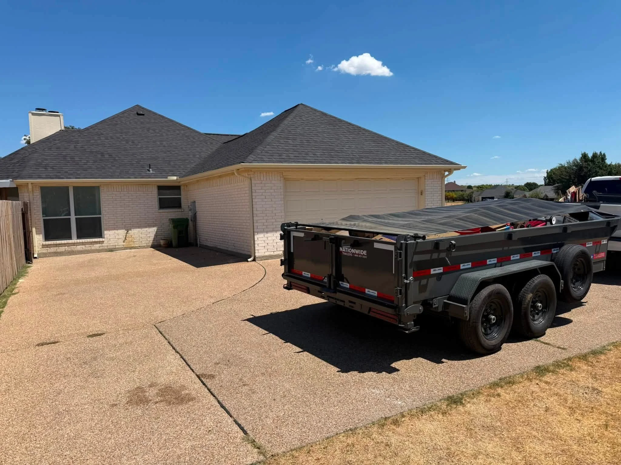 Backyard scene with a brick house, a garage with a sloped roof, and a black trailer in the driveway. The sky is clear with a few small clouds.