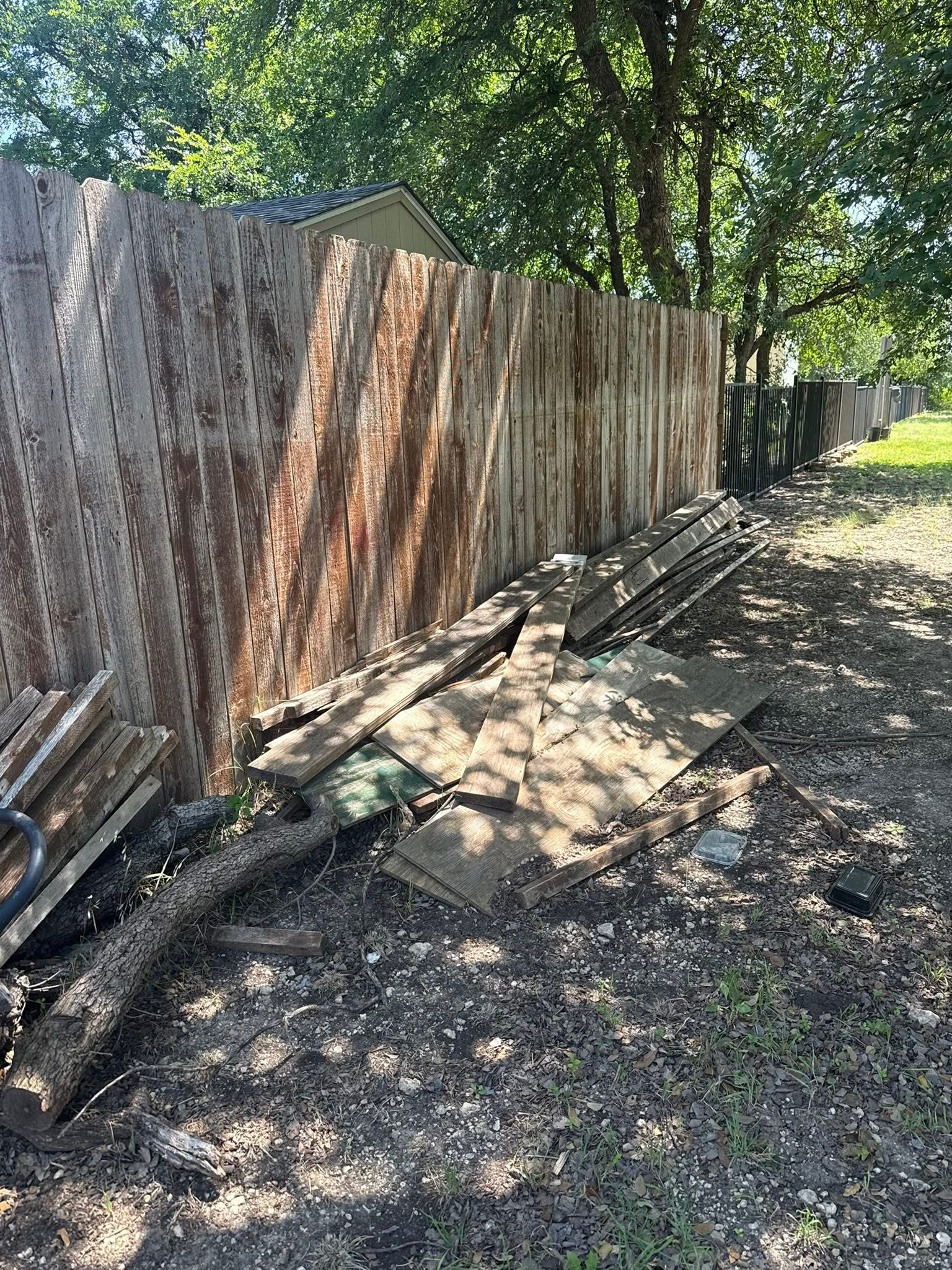 A wooden fence with some planks leaning on it, surrounded by dirt, roots, and small plants. There are trees and a house roof visible in the background on a sunny day.