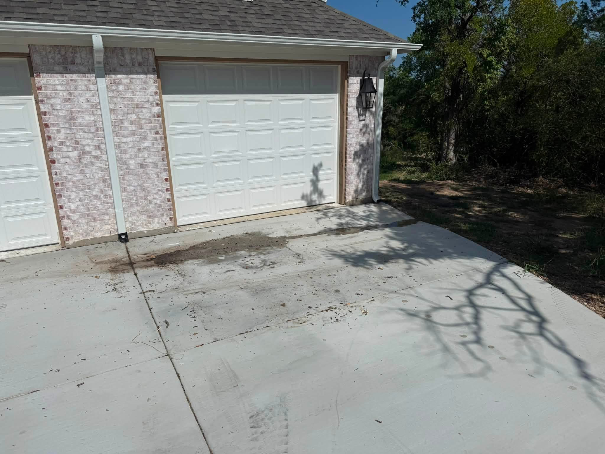 View of a white garage door on a house with brick exterior, a black outdoor lantern light next to it, and a concrete driveway with some dirt and shadows, surrounded by trees and clear blue sky.
