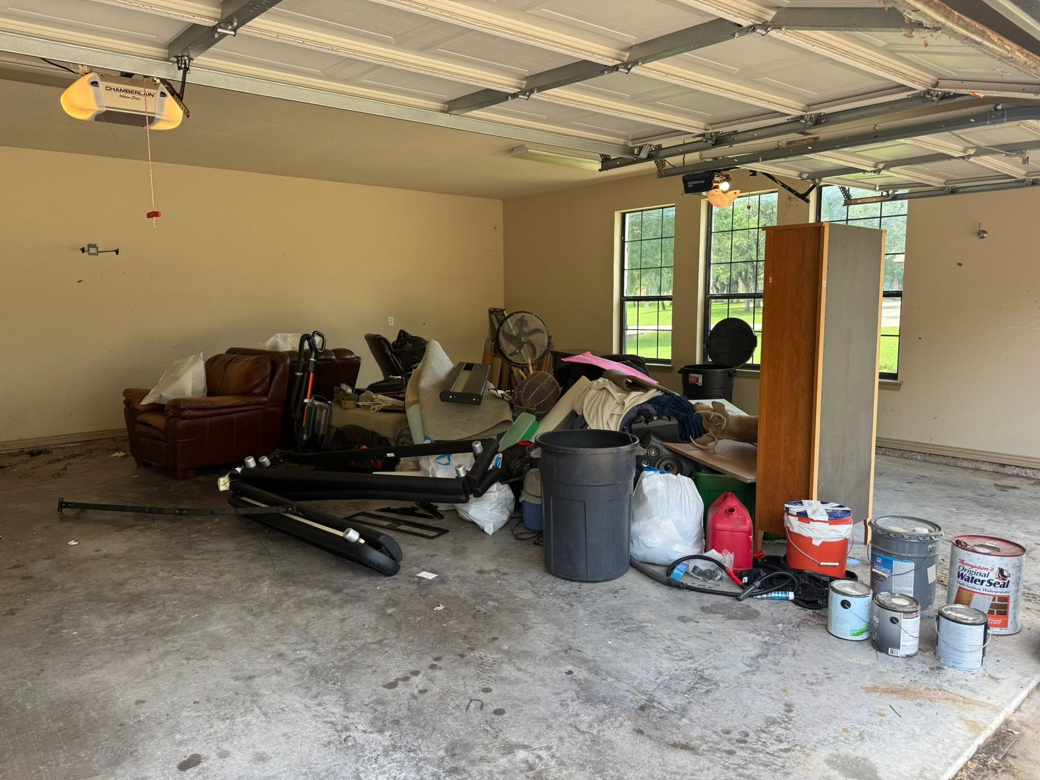 A cluttered garage with various household items, paint cans, and trash bins, and a brown leather couch near the wall, with large windows in the background.