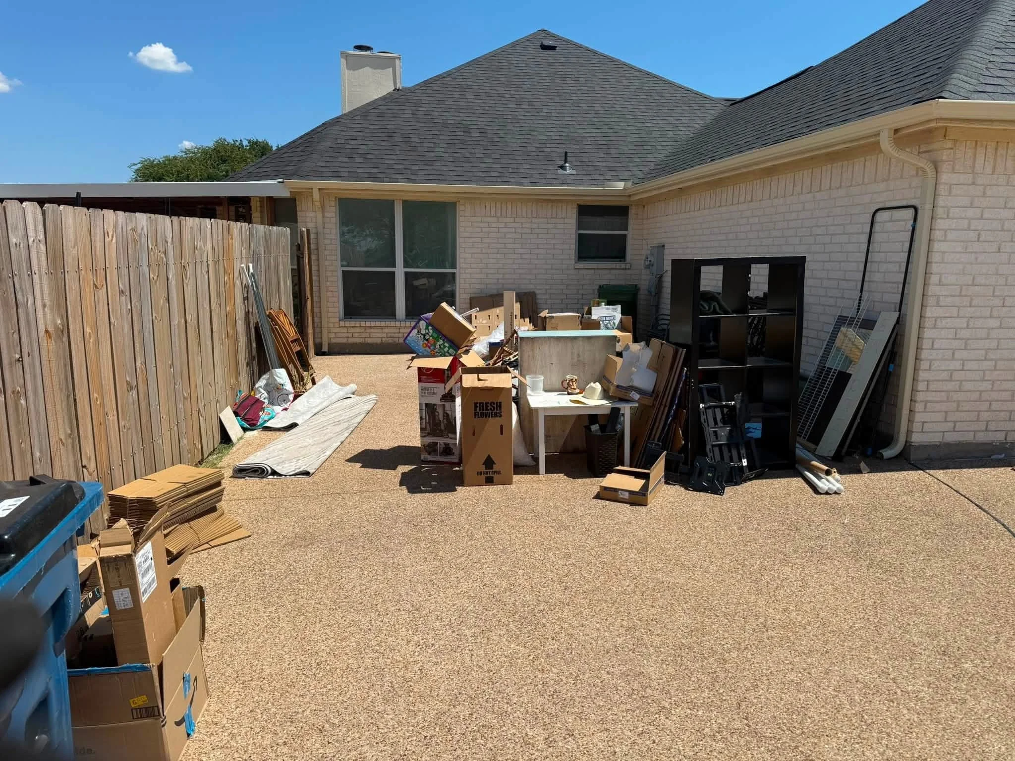 Backyard with pile of cardboard boxes and various items, wooden fence on the left, house with brick exterior and a gray shingle roof, clear blue sky.