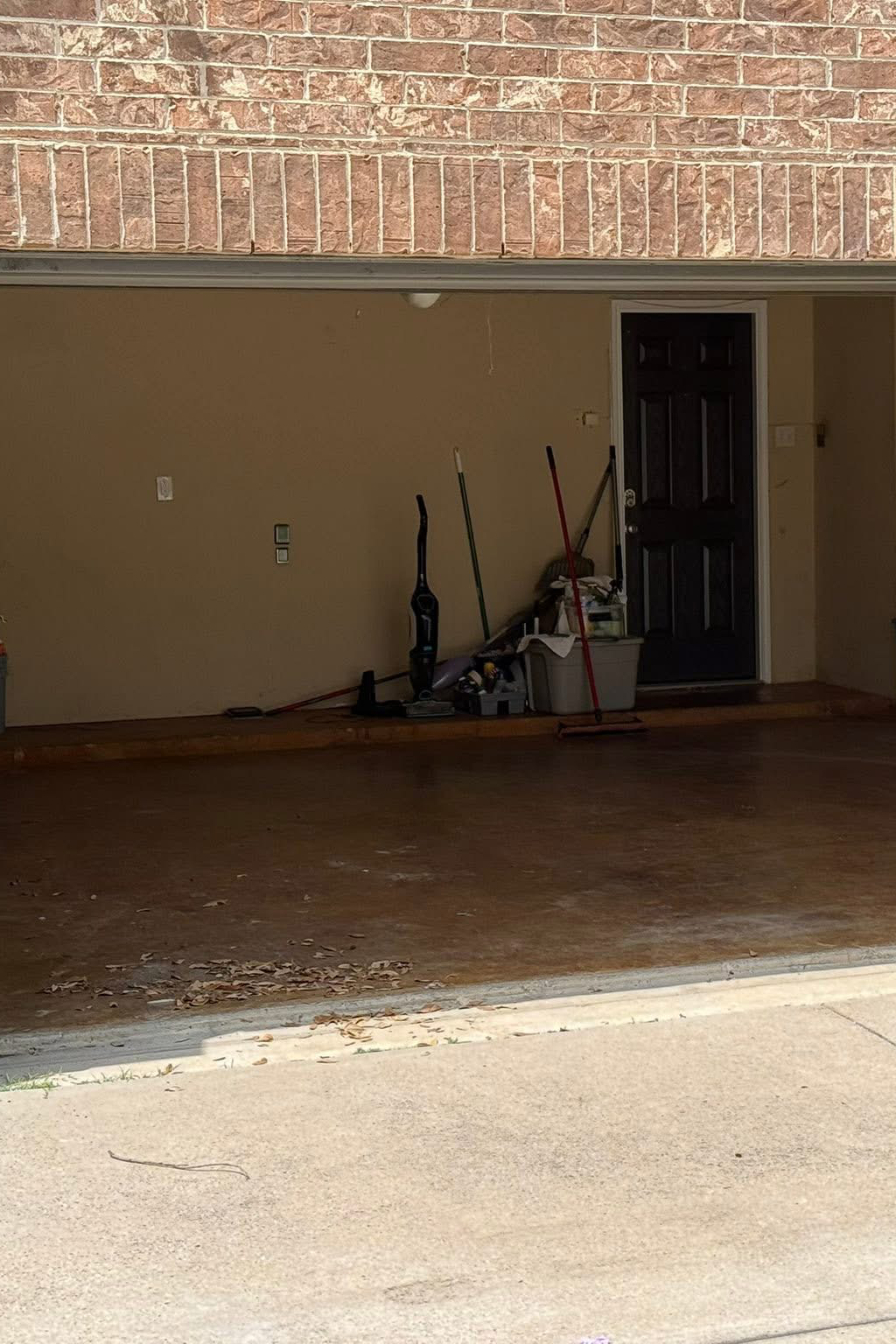 The garage interior with cleaning tools, a storage bin, and miscellaneous items near a door, with a brick exterior wall above.
