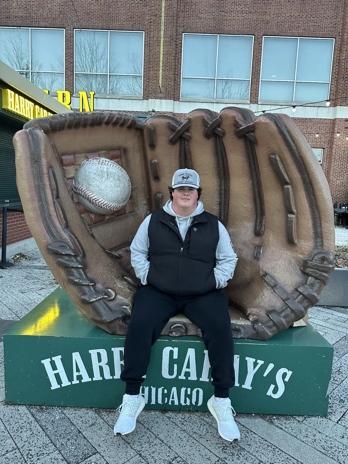 A young man sitting on a bench in front of a large softball glove and ball statue at Harry Caray's restaurant in Chicago.