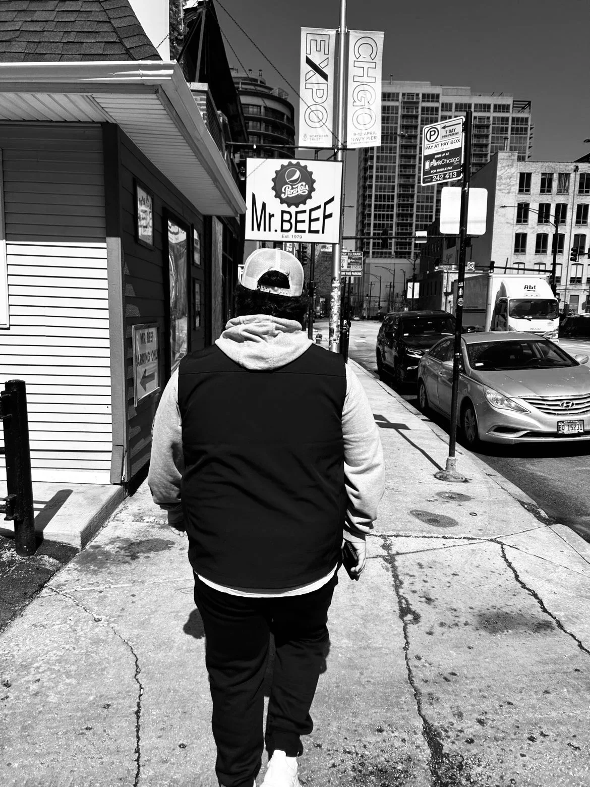 A person walking down a city sidewalk, wearing a baseball cap backwards, a hoodie, and a vest. Buildings, parked cars, and street signs are visible in the background in black and white.