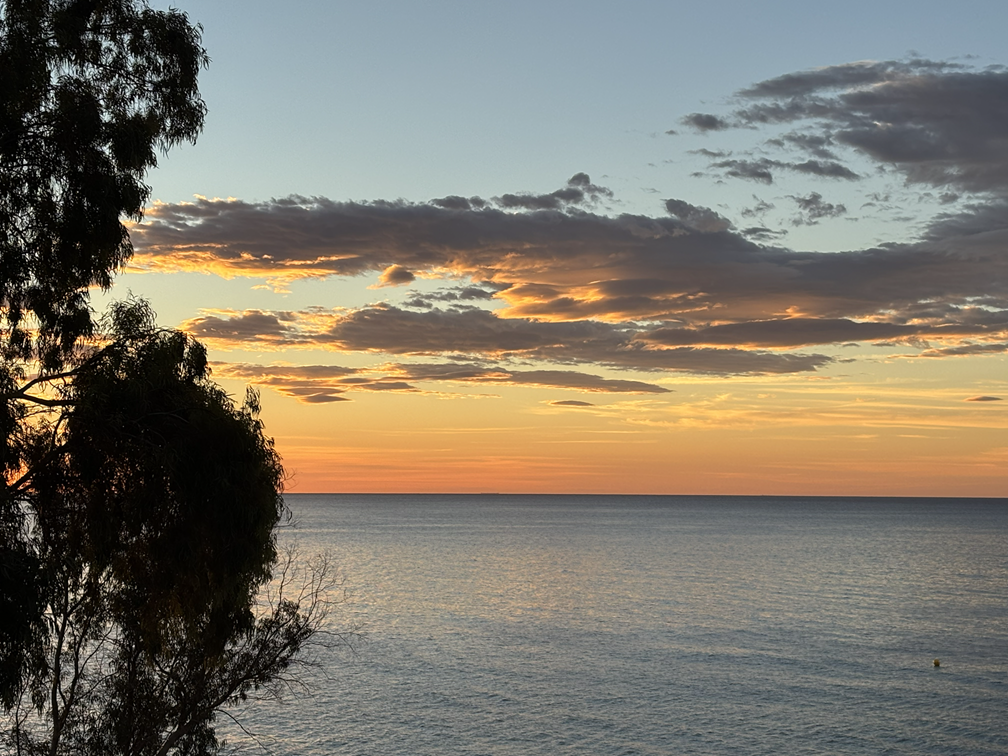 Sunrise over the Mediterranean Sea in Alicante, Spain, featuring calm waters, a sandy beach, rocky shoreline, and a colorful morning sky.