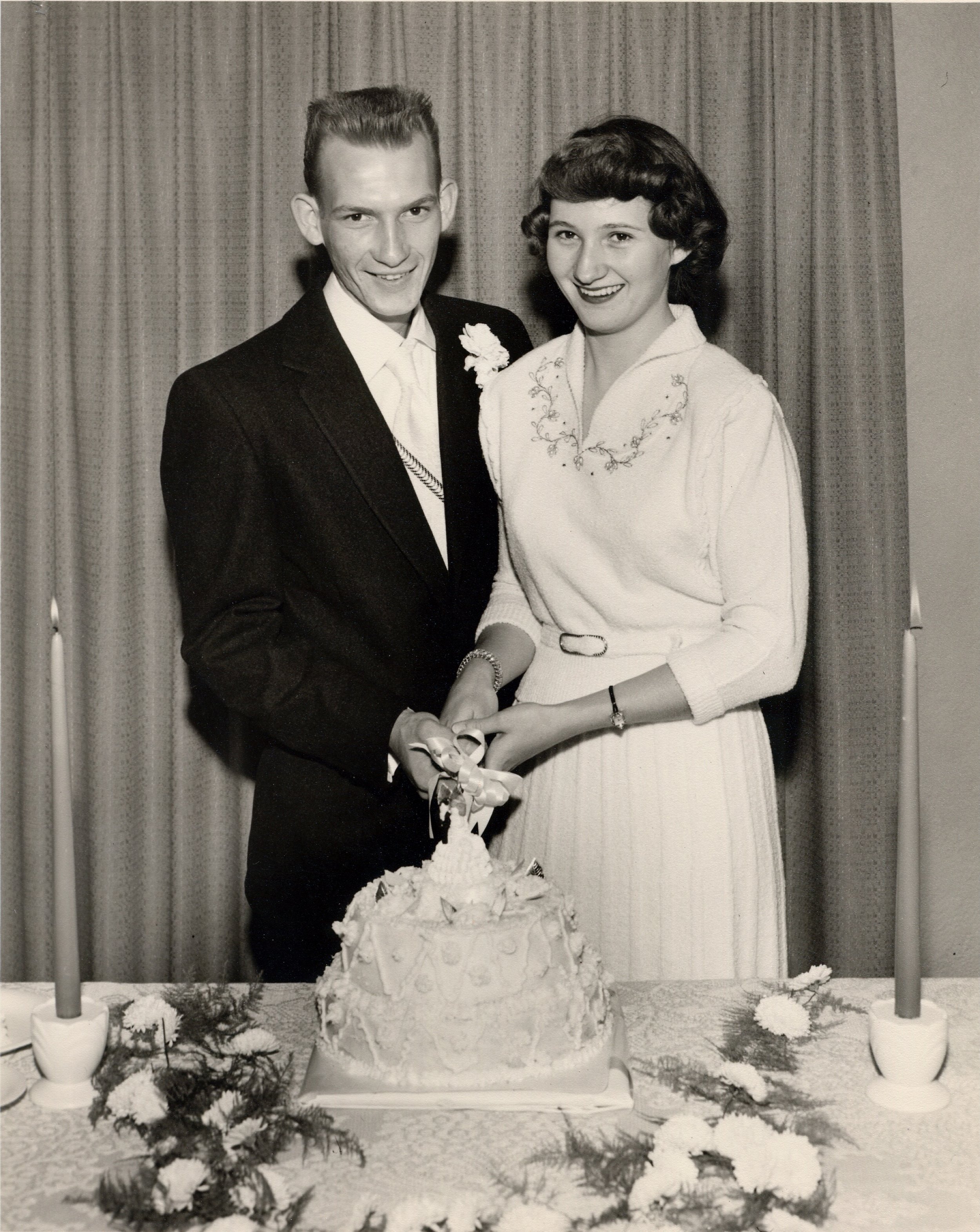 Black and white photograph of a young couple at a wedding, standing behind a wedding cake with a floral and ribbon decoration. The man is in a dark suit with a white shirt and a flower boutonniere, and the woman is in a light-colored blouse with embroidery, smiling and holding hands with the man. The background has curtains, and there are candles and decorative greenery on the table with the cake.