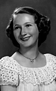 Black and white portrait of a smiling young woman with wavy hair, wearing a lace dress and a necklace. This is my Mom, Gloria Sumner